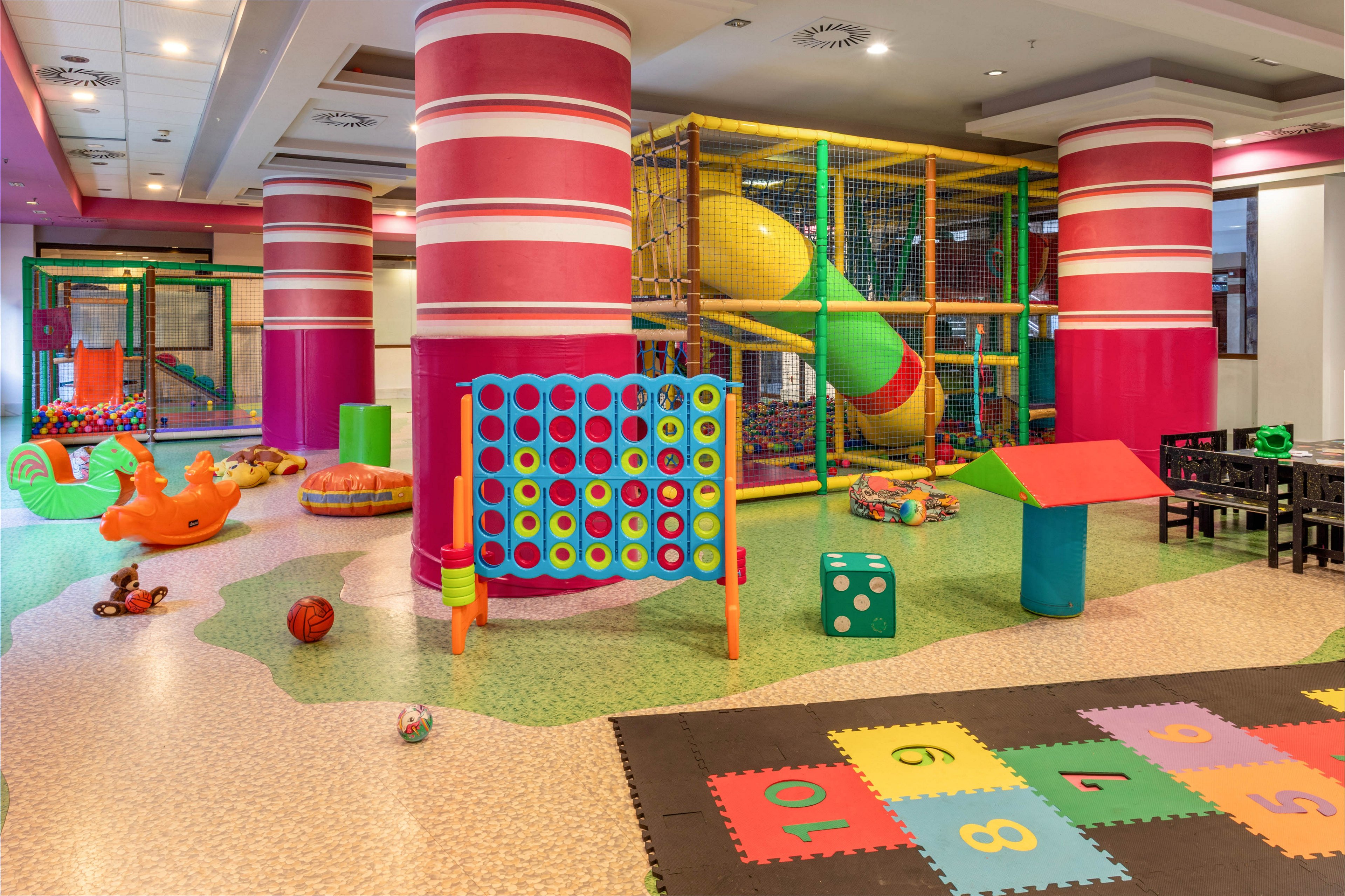 Brightly colored indoor playroom featuring large pink columns and multiple play structures. A giant connect-four style game stands in the center alongside soft blocks and scattered balls. In the background, a multi-level climbing frame with yellow tube slide and ball pit is enclosed by safety netting. A hopscotch mat with numbers 1 through 10 adds educational play to the cheerful, family-friendly environment.