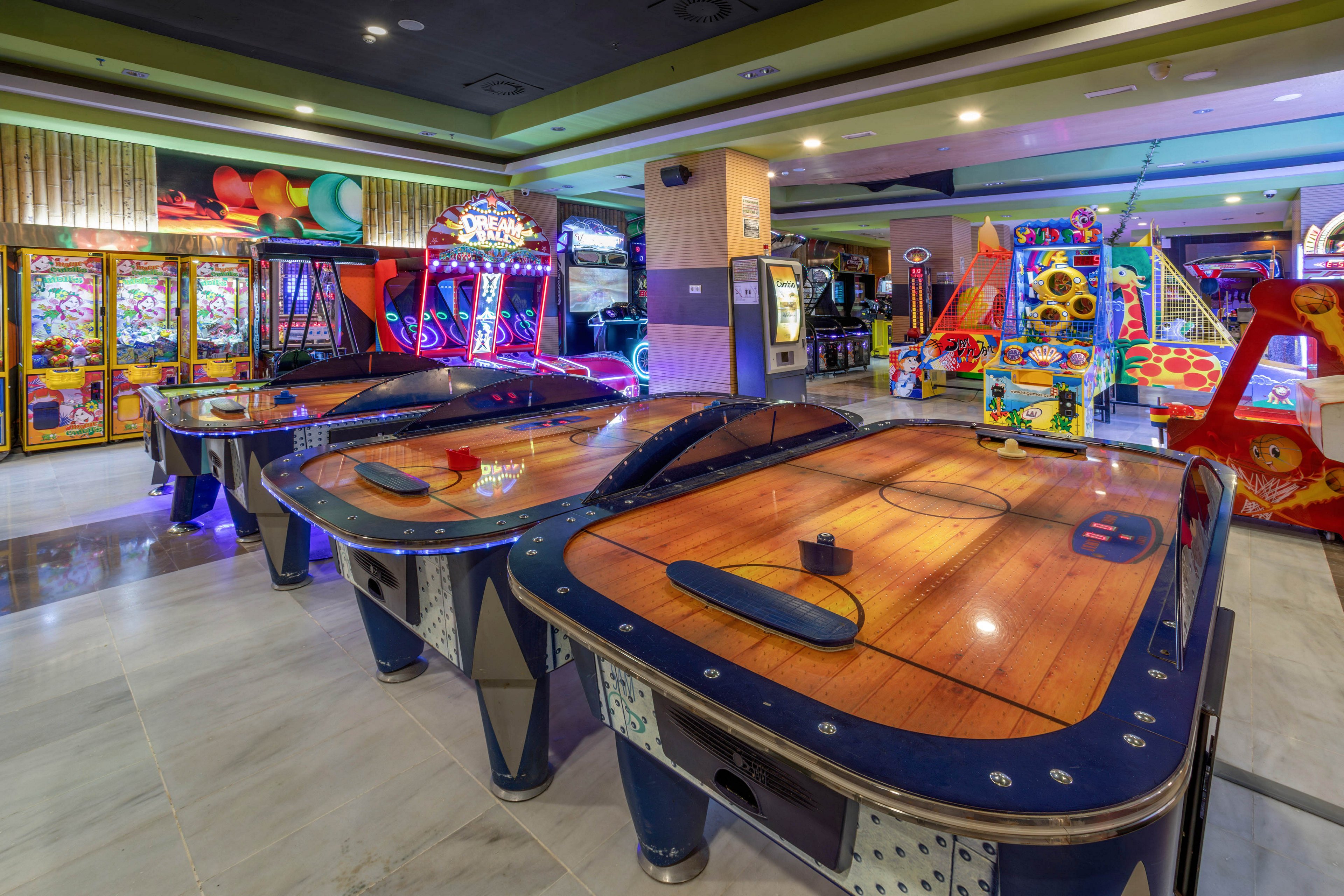 A row of air hockey tables stands in the foreground of a brightly lit indoor arcade. Various electronic game machines with vivid lights and colorful graphics line the walls in the background. The polished floor reflects the neon and LED glows, adding to the energetic atmosphere. The scene appears ready for players, but no people are currently present.