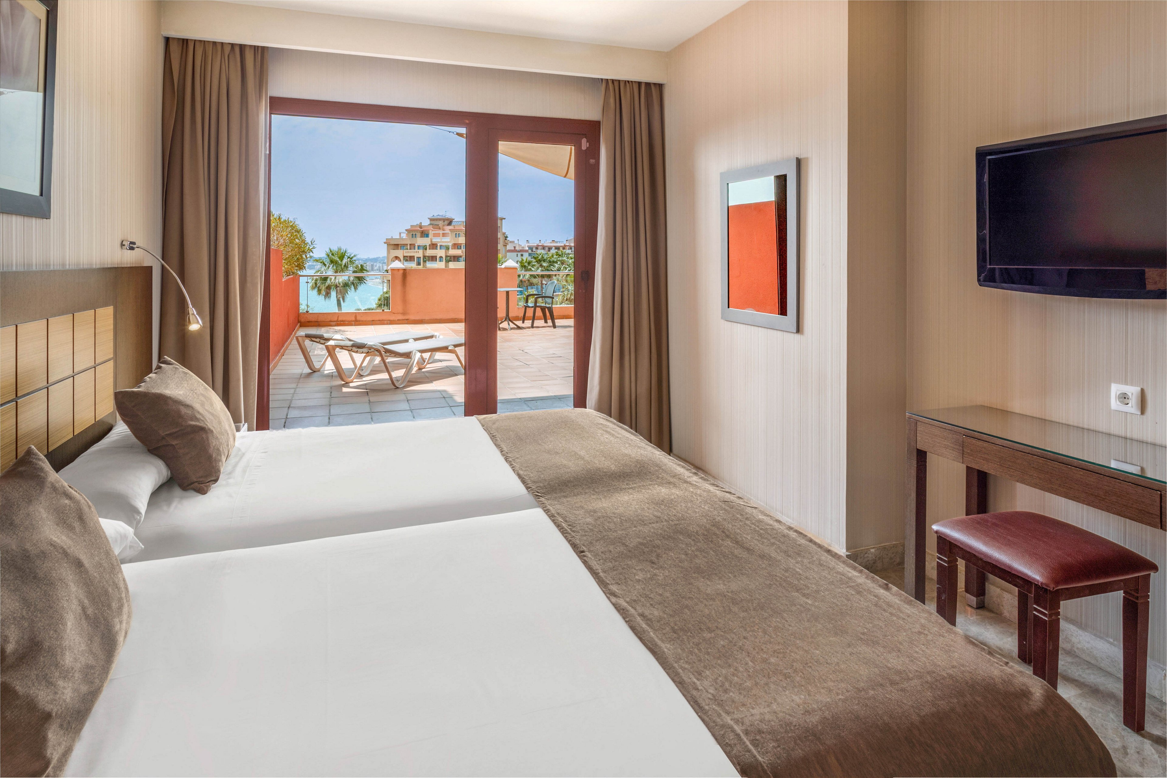A contemporary hotel room features two neatly made beds with neutral bedding and cushions. Large glass doors open onto a private terrace with sun loungers and a view of the sea and nearby buildings. The interior includes a wall-mounted flat-screen TV, a small desk, and a stool, all in warm, earthy tones. Natural light fills the space, creating a calm and inviting atmosphere for guests.