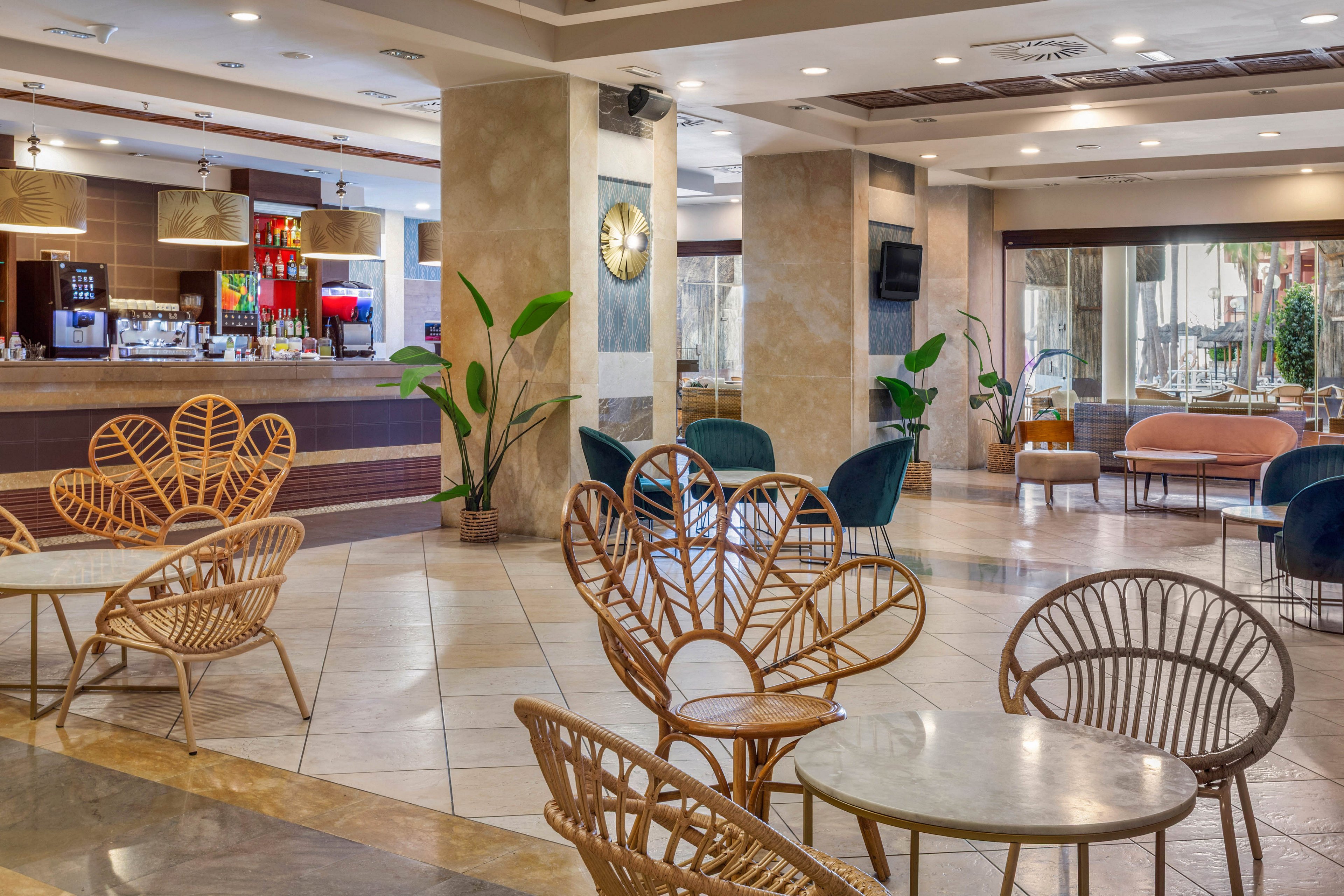 Spacious hotel lobby lounge featuring stylish rattan chairs and round tables. A contemporary bar counter with illuminated shelves and drink displays is visible in the background. Large marble columns, tiled flooring, and indoor plants create a clean, modern atmosphere. Soft seating areas with armchairs and sofas provide comfortable spaces for guests to relax or socialize.