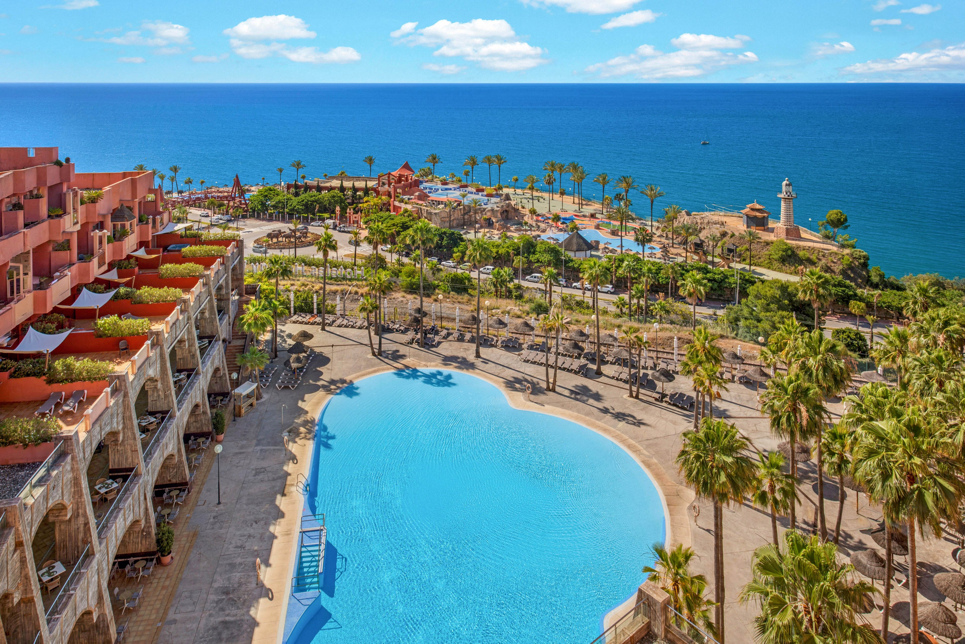 A large, bright blue swimming pool sits at the center of a seaside resort overlooking the ocean. Terraced hotel buildings with balconies line one side, while tall palm trees surround the pool and pathways. In the distance, additional resort structures and a small lighthouse-like tower appear near the coastline. The sky is clear with scattered white clouds, creating a sunny, inviting vacation atmosphere.