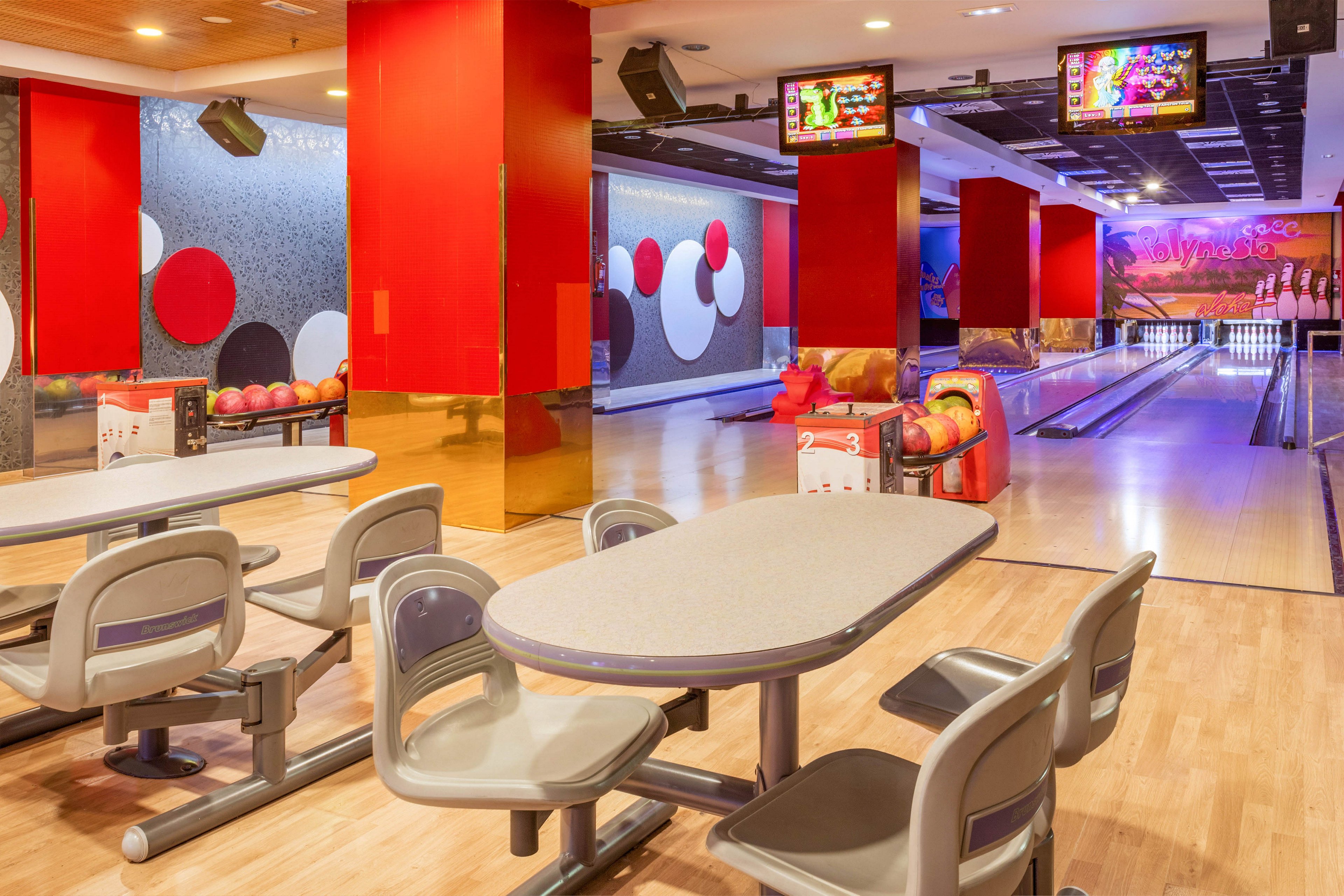 A brightly lit bowling alley features several empty lanes and ball returns. Modern gray seating and tables are arranged in the foreground on a polished wooden floor. Vibrant red columns and colorful wall graphics with circular patterns add energy to the space. Overhead screens display bowling graphics, creating a lively entertainment atmosphere despite the absence of people.