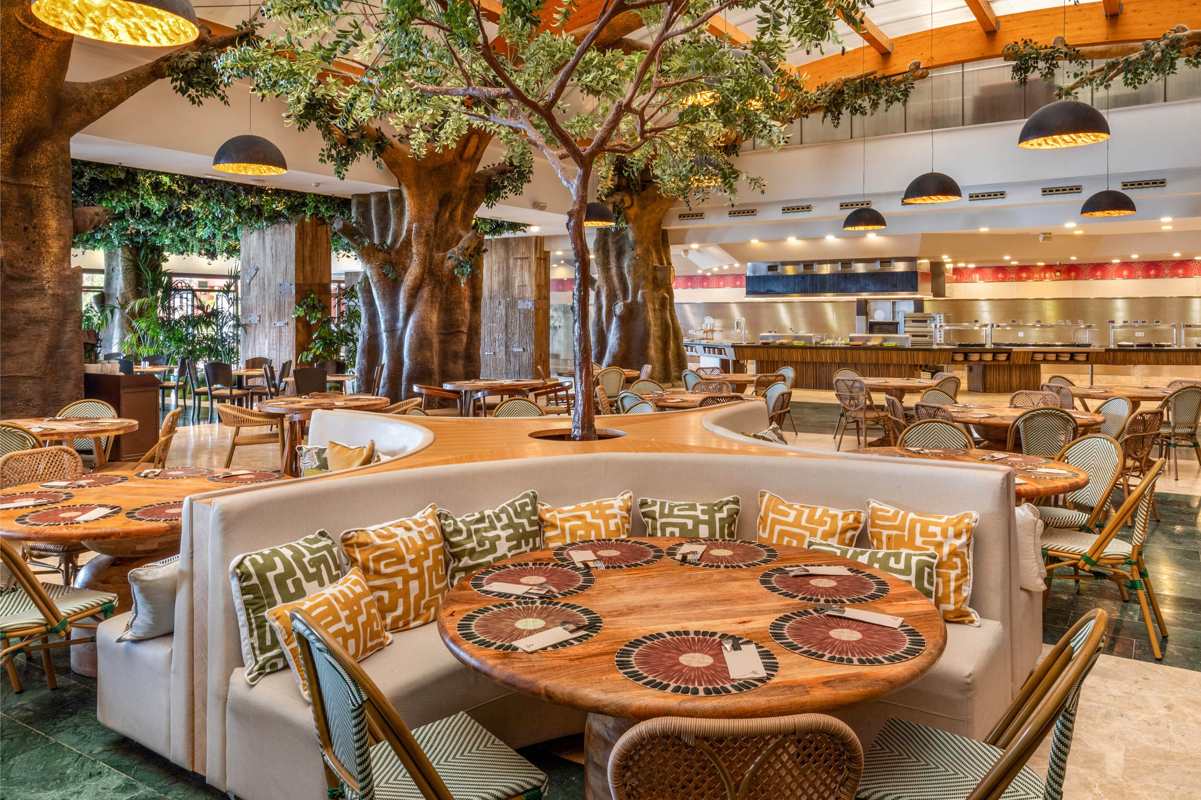 A spacious contemporary restaurant features round wooden tables surrounded by cushioned banquette seating and woven chairs. Large artificial trees with lush green foliage rise through the floor, creating a natural, indoor garden atmosphere. Warm pendant lights hang from a wooden ceiling, illuminating patterned pillows and decorative tabletops. In the background, an open kitchen and service counter add to the lively, welcoming dining environment.