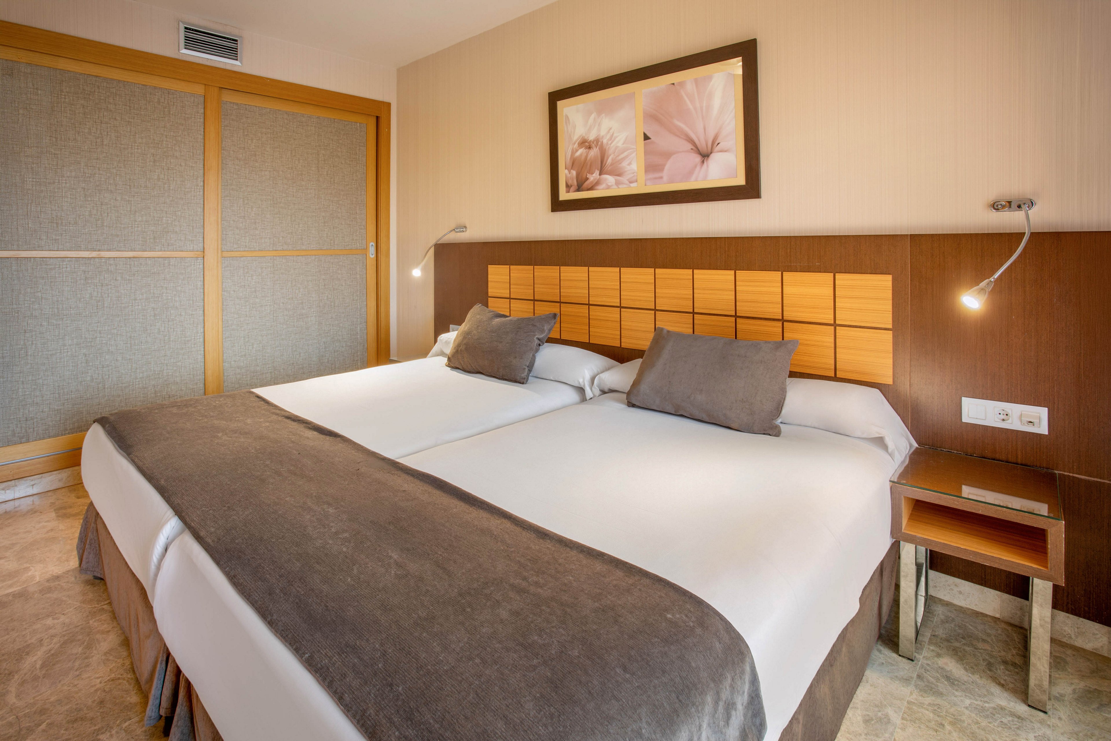 A contemporary hotel room features two neatly made twin beds pushed together with neutral bedding and accent pillows. The headboard combines wood and upholstered panels, complemented by built-in reading lights on each side. A framed floral artwork hangs above the bed, adding a soft decorative touch. Sliding closet doors and a small bedside table complete the clean, minimalist interior design.