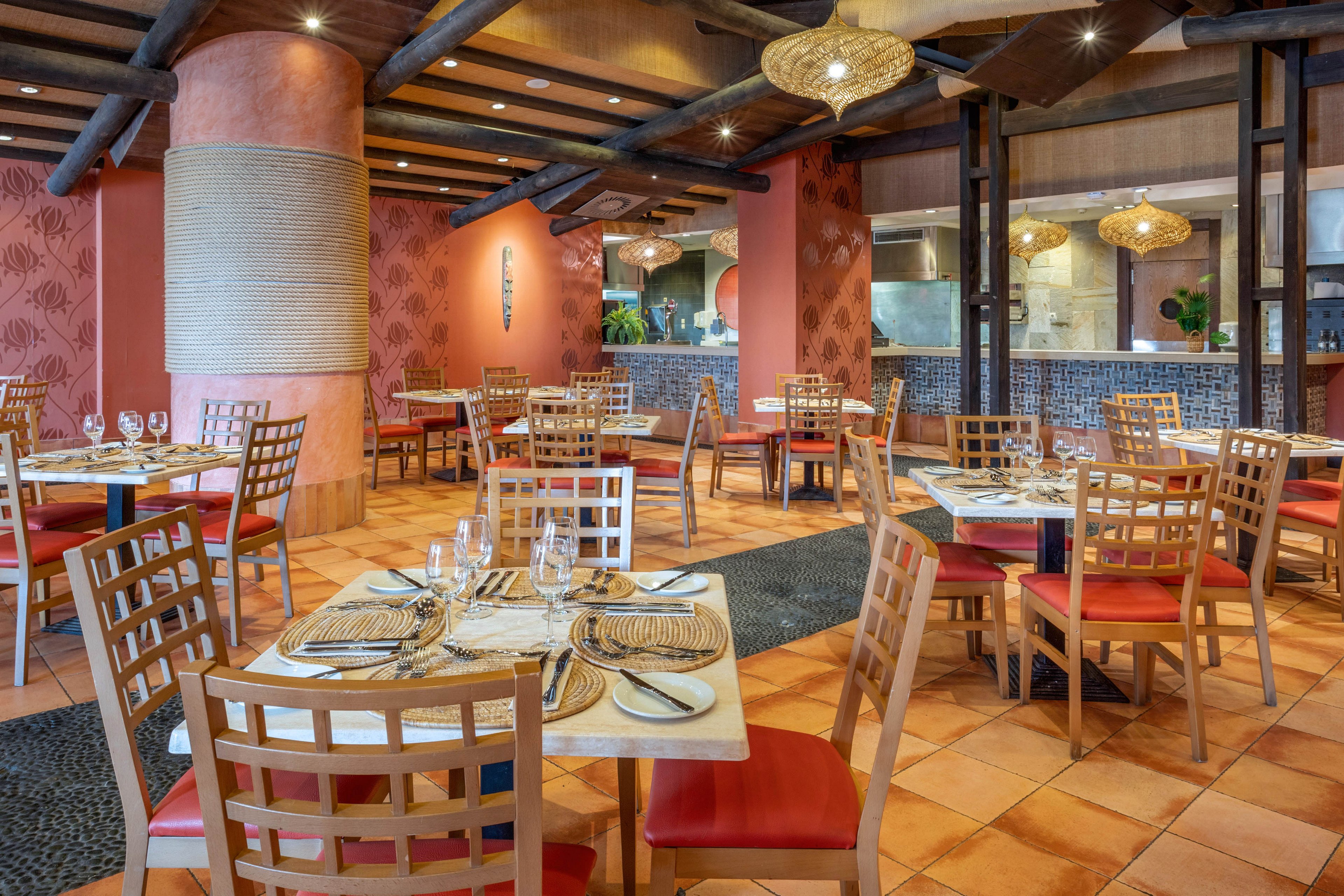 A warmly lit restaurant dining room features neatly set tables with woven placemats, glassware, and cutlery. Wooden chairs with red cushions surround square tables arranged across a tiled floor. Earthy tones, textured columns, and patterned walls create a Mediterranean or tropical ambiance. Hanging wicker light fixtures and an open kitchen area in the background add to the inviting, casual dining atmosphere.