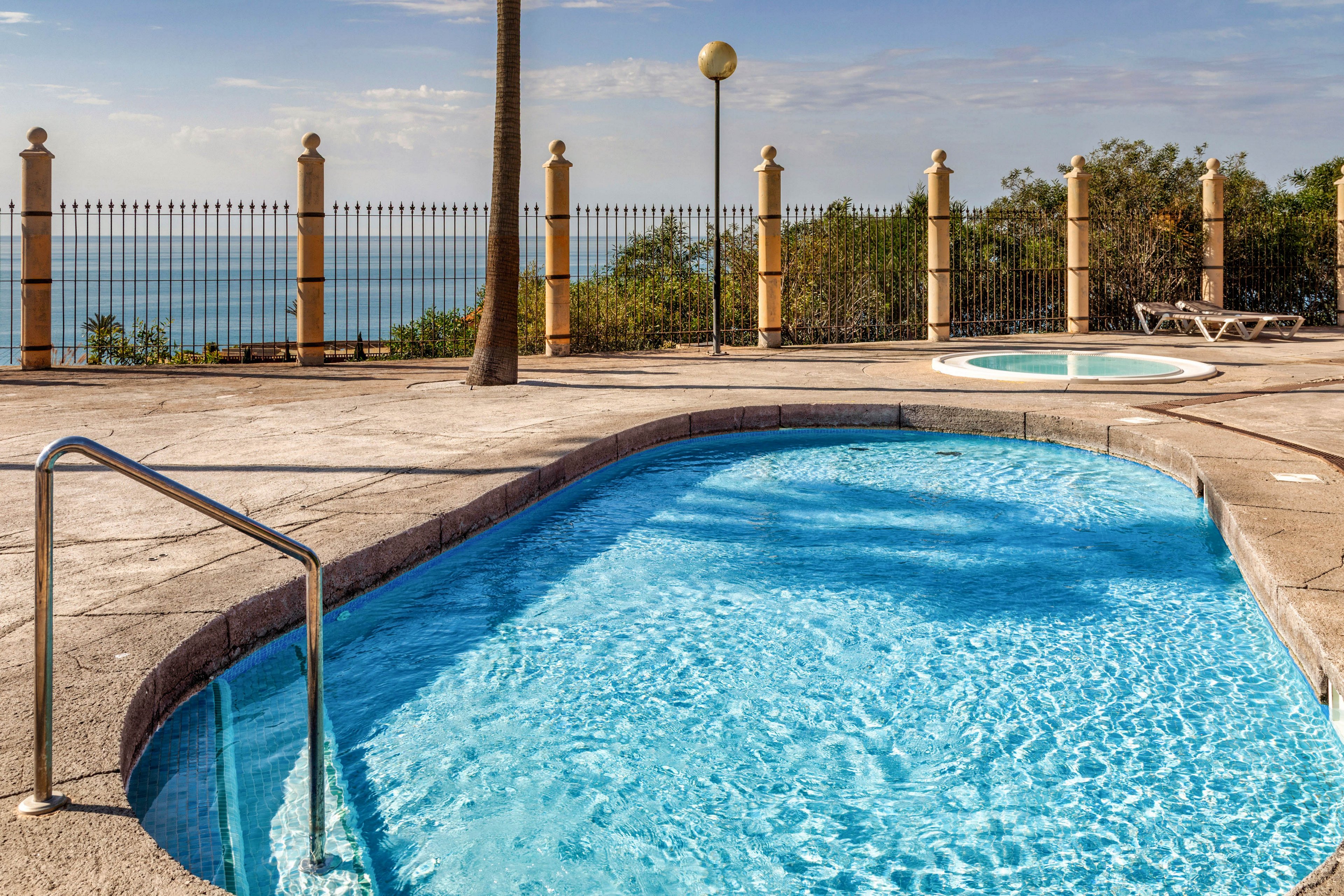An outdoor swimming pool with clear blue water sits on a sunlit terrace. A metal handrail leads into the pool, with a small circular hot tub visible in the background. The area is surrounded by a decorative fence and tall posts, opening to a scenic view of the ocean and distant horizon. The bright sky and calm water create a relaxed, vacation-like atmosphere.