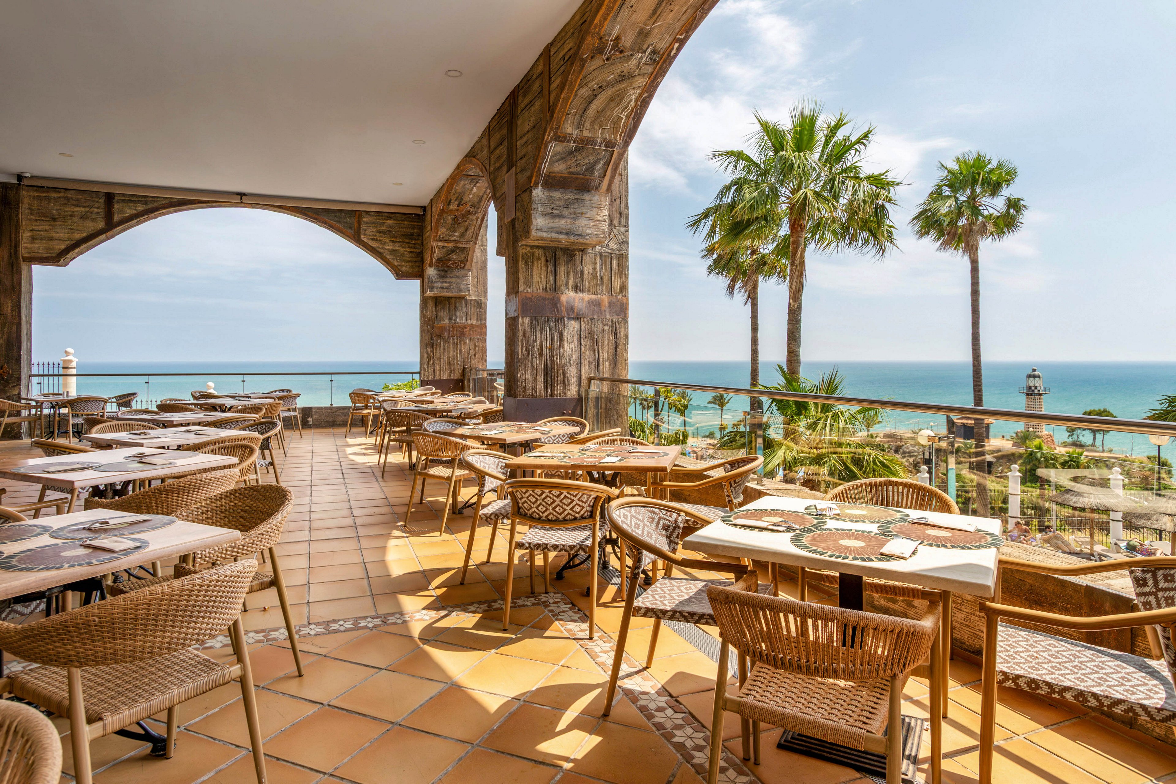 An open-air restaurant terrace overlooks a bright blue sea on a sunny day. Wicker chairs and round tables are neatly arranged on a tiled floor beneath large stone arches. Palm trees and coastal landscaping frame the view toward the water and distant horizon. The scene feels relaxed and inviting, ideal for outdoor dining by the ocean.