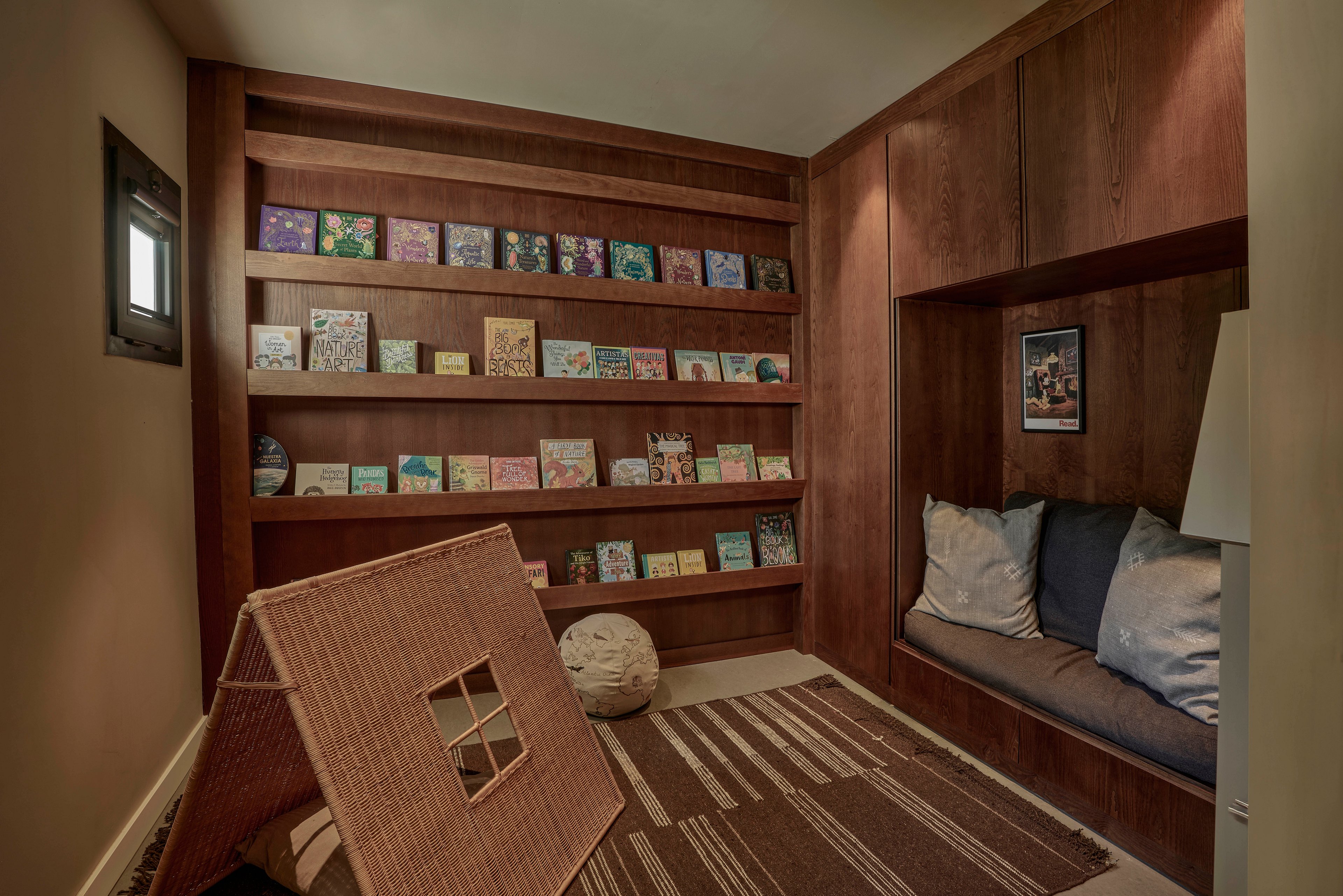 A small, cozy room features a built-in wooden bookcase filled with colorful children's books. A cushioned bench with large pillows is set into the wood paneling, creating a comfortable reading nook. On the floor, a patterned rug and a woven play structure add warmth and texture to the space. Soft lighting and neutral walls contribute to a calm, inviting atmosphere ideal for quiet reading or relaxation.
