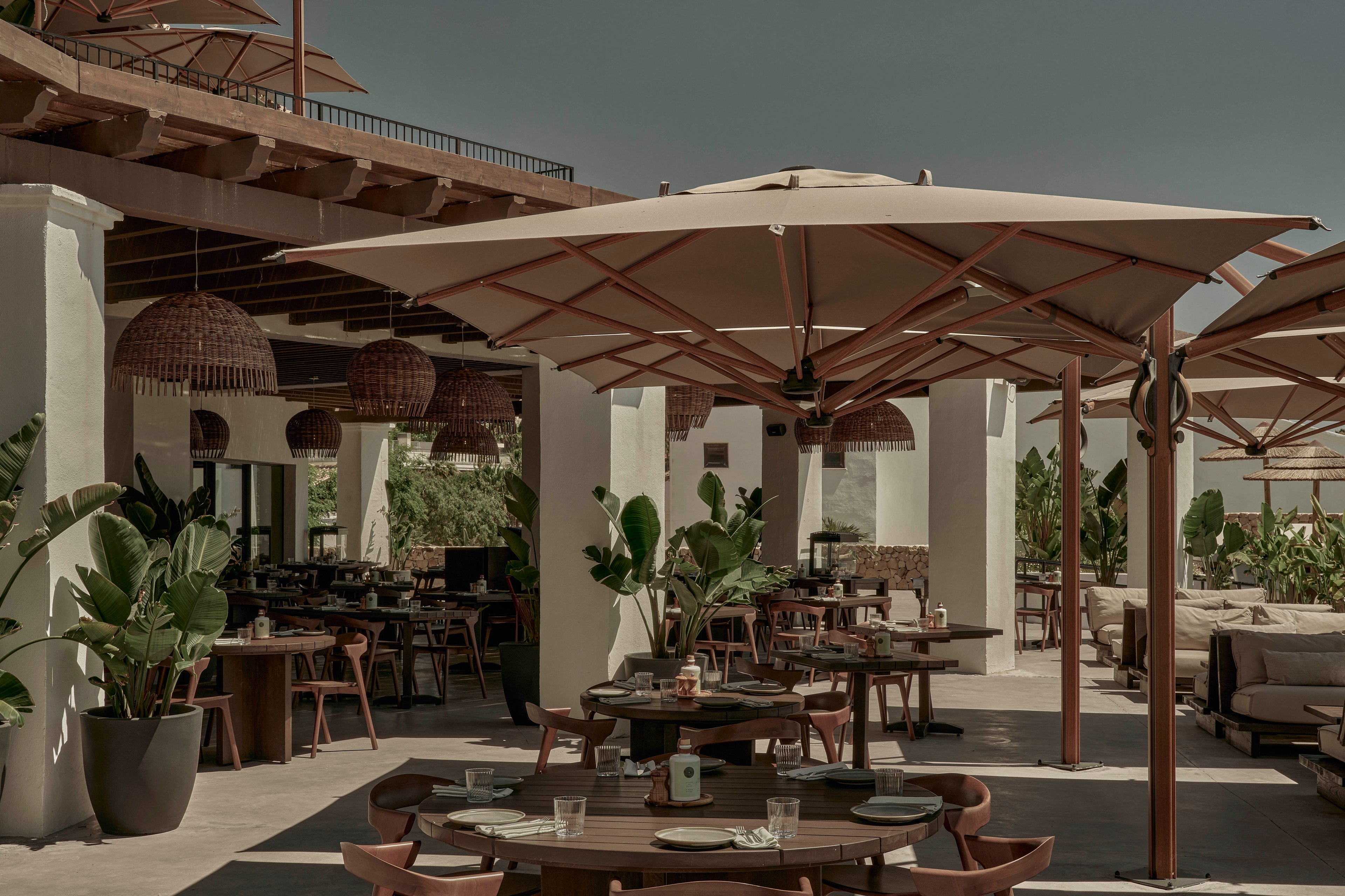 An elegant open-air restaurant terrace features round wooden tables set with plates, glasses, and napkins. Large beige umbrellas and a wooden pergola provide shade over the seating area. Lush potted plants add greenery between the tables and along the edges of the space. The muted, warm color palette and soft lighting create a relaxed, sophisticated atmosphere.
