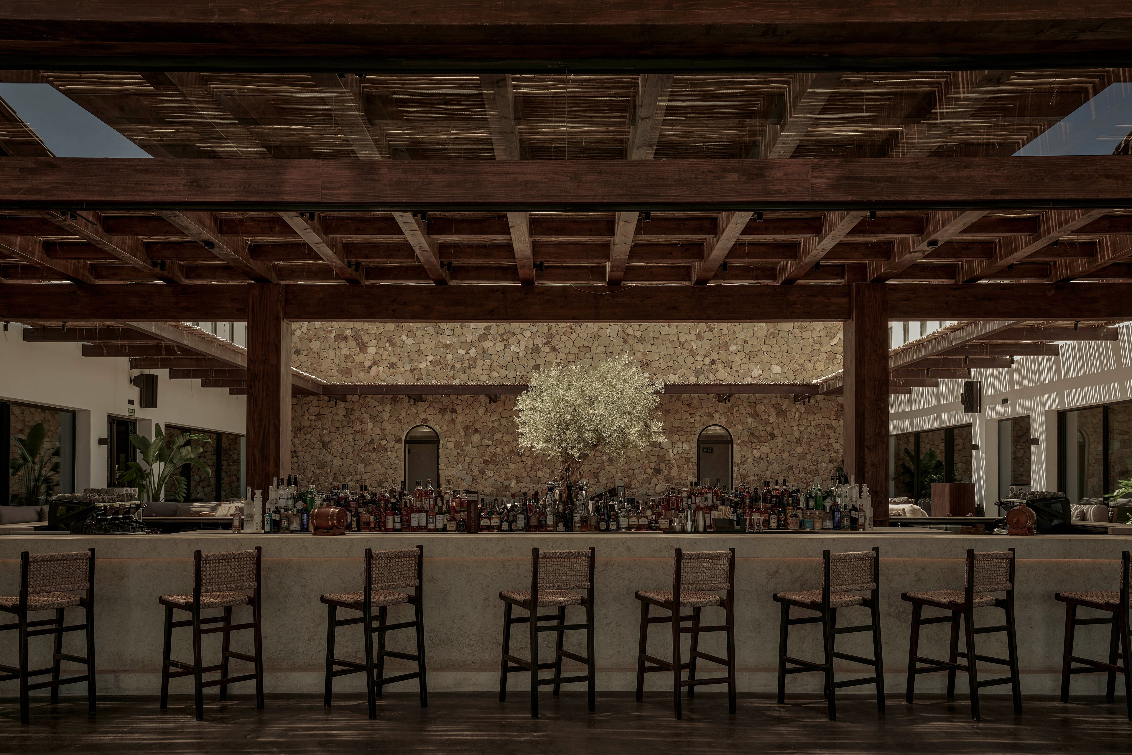 An open-air bar with a long concrete counter and wooden stools is set beneath a large wooden pergola. Bottles and glassware line the back of the bar, creating a focal point in the center of the space. A stone wall with arched niches and a small tree adds texture and warmth to the background. The muted, earthy color palette and soft lighting give the scene a calm, sophisticated atmosphere.