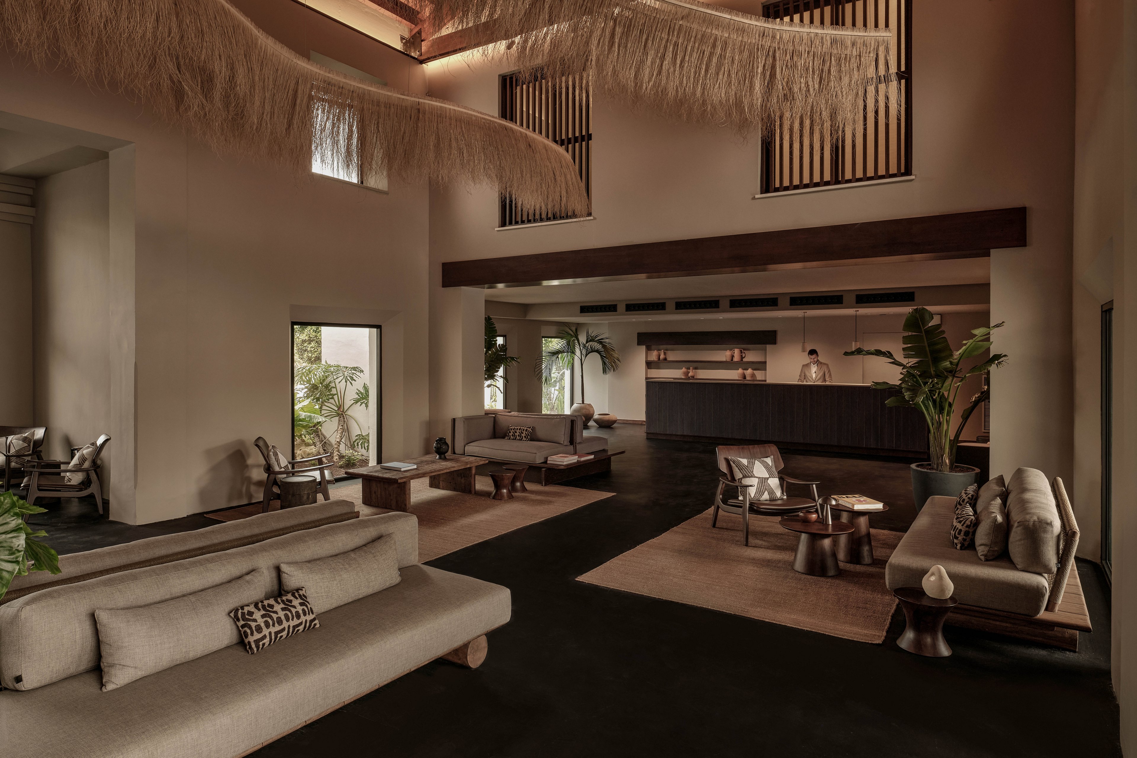 Spacious contemporary lounge features low-profile sofas and warm neutral tones. The setting includes an open bar at the back where a person stands, with seating areas arranged on large rugs. Tall indoor plants, textured ceiling elements, and high windows add depth and natural light. Overall, the space feels calm, sophisticated, and inviting with a minimalist design aesthetic.