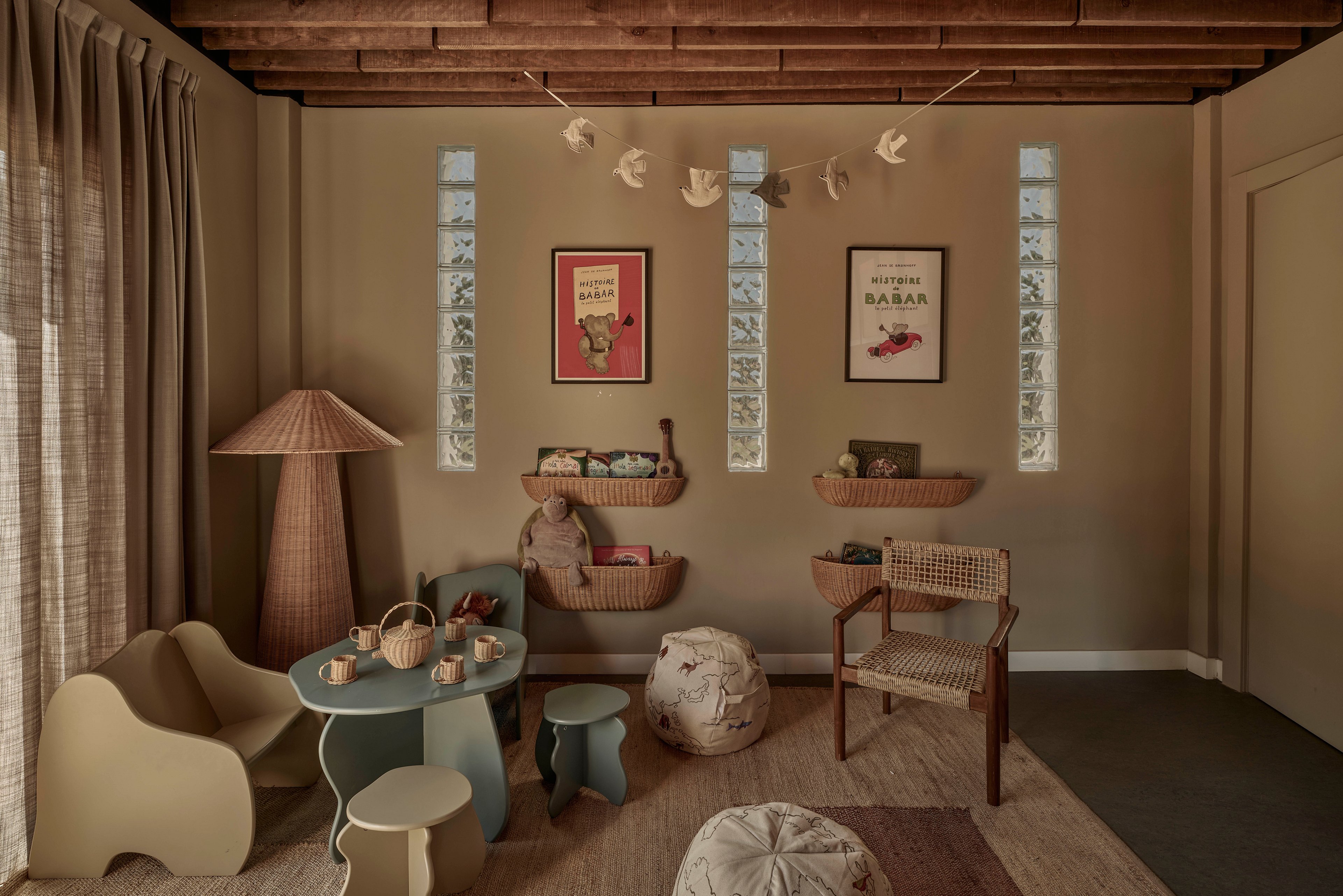 A warm, neutral-toned children's playroom features a small table with matching stools and various toys arranged neatly. Wicker baskets mounted on the wall hold stuffed animals and books, while a woven chair and floor cushions add comfort. Soft curtains filter light from a large window, and decorative prints and hanging birds accent the walls. The overall scene feels calm, organized, and inviting for quiet playtime.