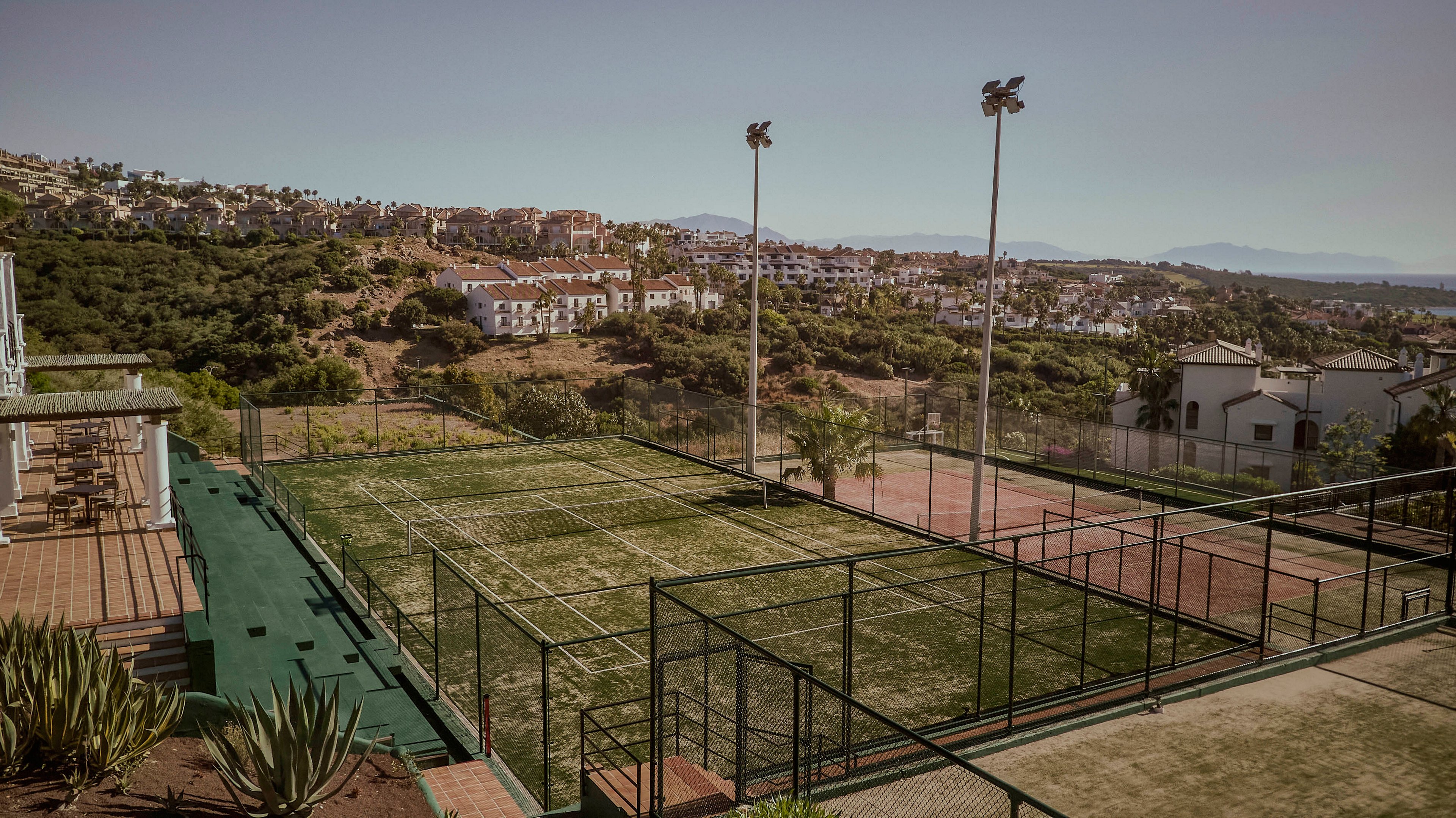 An outdoor sports facility with fenced courts sits on a hillside overlooking a sprawling residential area. The foreground shows a worn green court surface beside a red court, bordered by railings and desert-style plants. Tall floodlights rise above the courts, and houses stretch across the distant slopes under a hazy sky. The overall scene feels quiet and sunlit, suggesting a warm, dry climate.