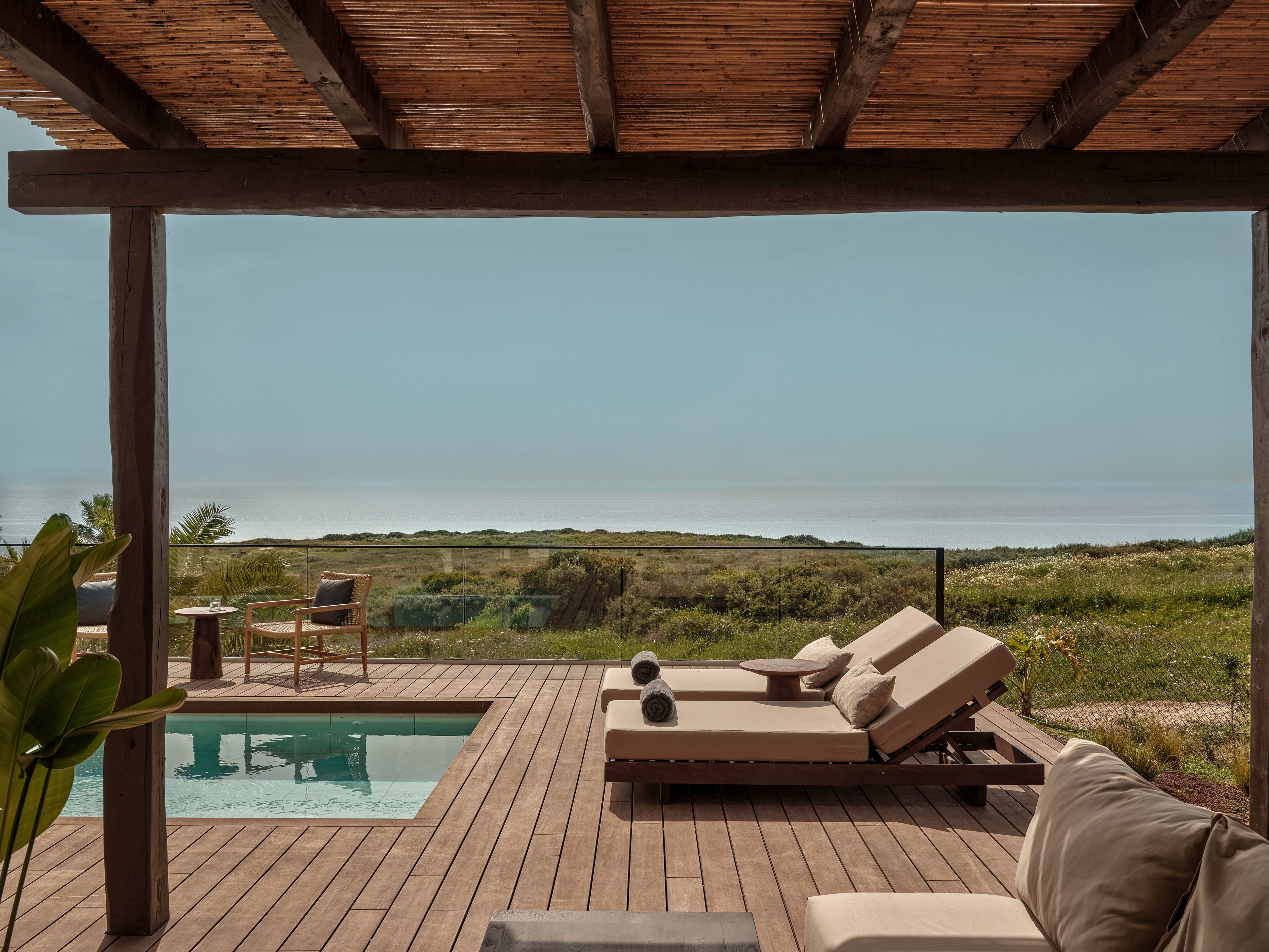 A serene outdoor deck features cushioned lounge chairs beside a small rectangular swimming pool. The space is shaded by a wooden pergola and framed by minimalist outdoor furniture. Beyond the deck, a grassy coastal landscape stretches toward the horizon under a clear sky. The scene emphasizes relaxation, natural materials, and a tranquil seaside atmosphere.