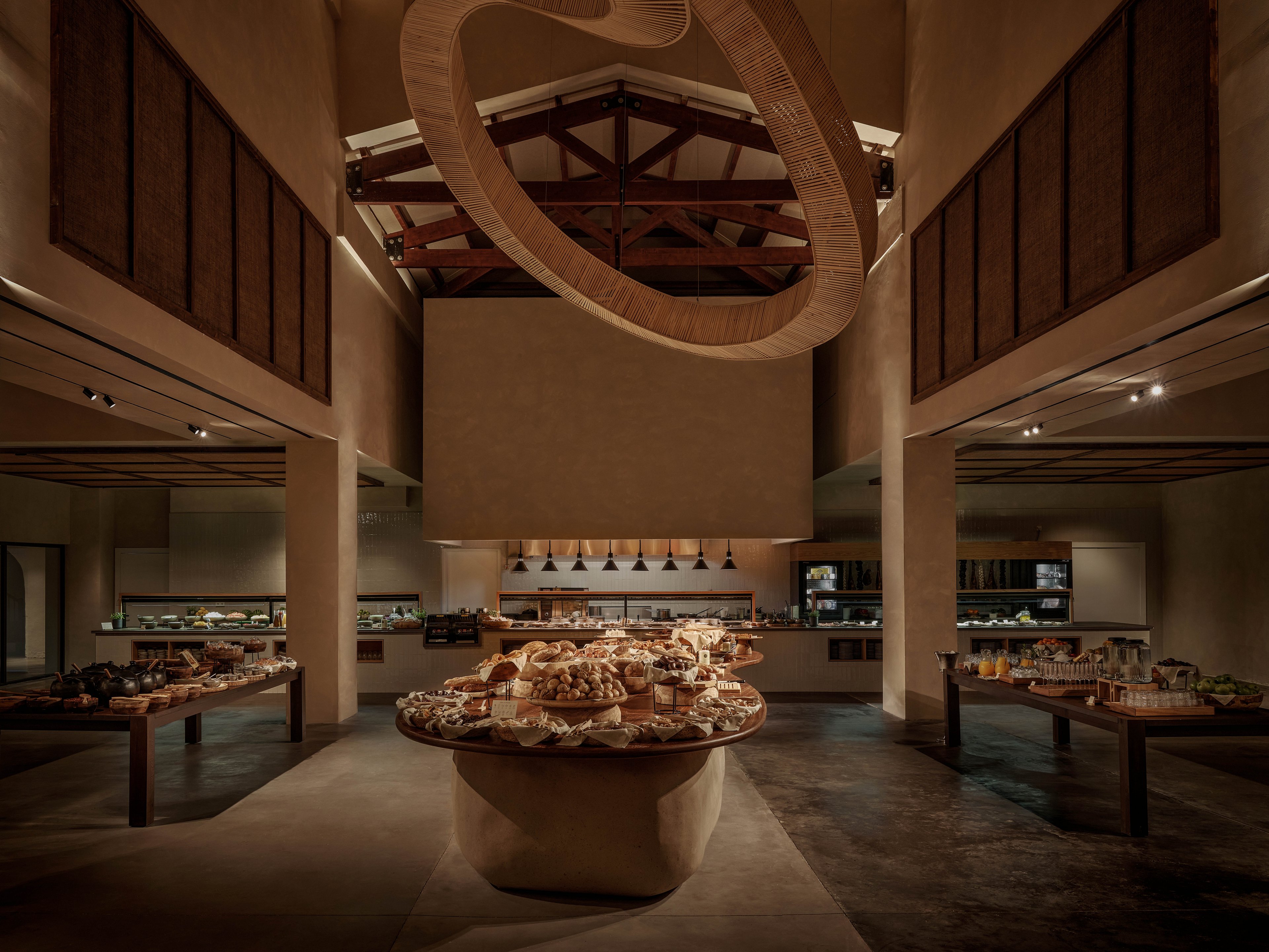A large circular buffet table filled with breads, pastries, and assorted dishes stands at the center of a spacious restaurant. The setting features warm lighting, high ceilings with exposed beams, and minimalist architectural details. Additional food stations and counters line the walls, showcasing more culinary options. The overall atmosphere feels refined and inviting, with a focus on gourmet dining and sophisticated interior design.