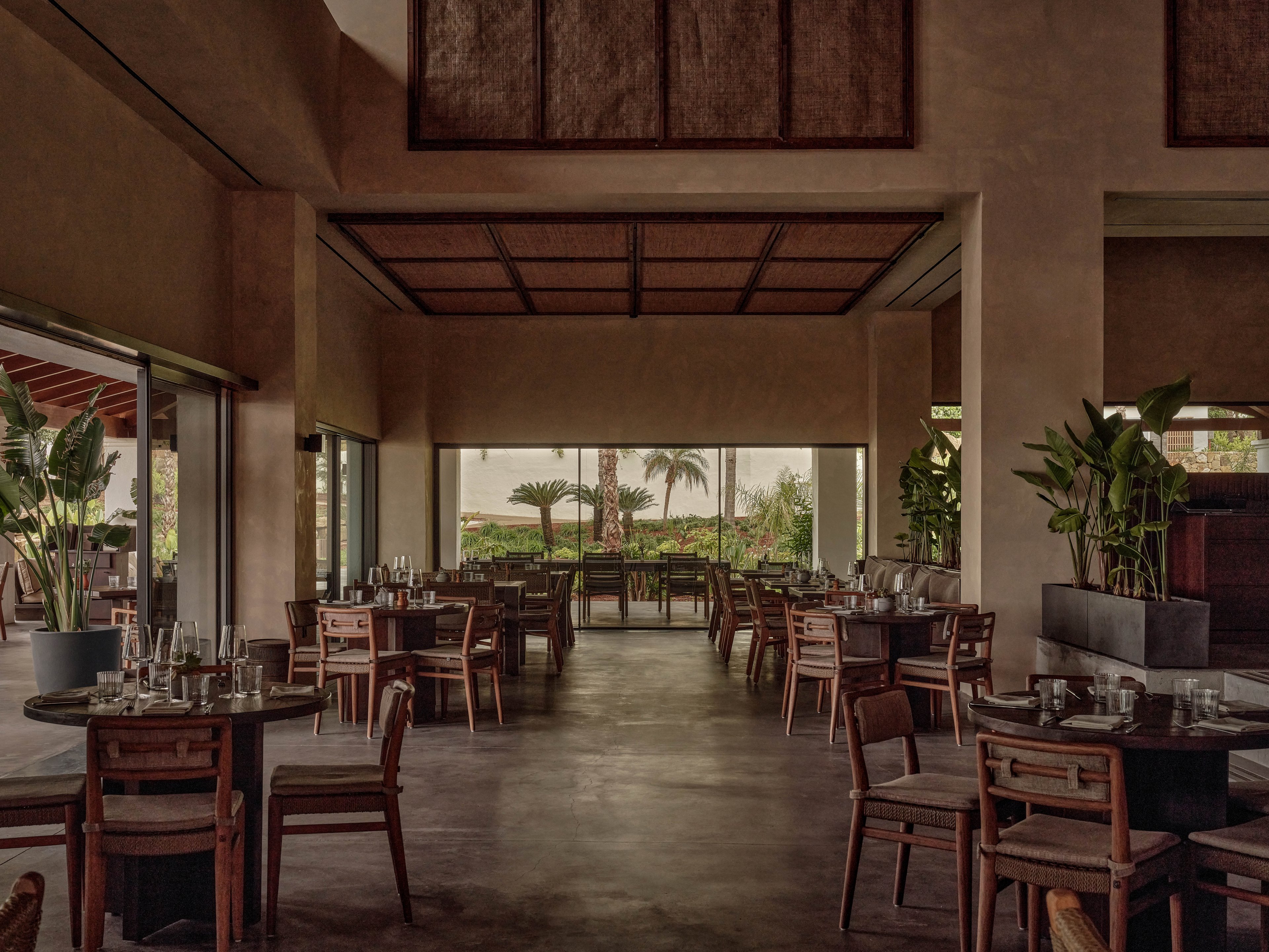 A spacious restaurant interior features wooden tables and chairs neatly arranged in an open-plan dining area. Large windows and sliding doors reveal a lush tropical garden with palm trees in the background. Soft, warm lighting and earthy tones create a calm, inviting atmosphere. Potted plants and minimalist decor enhance the natural, resort-like setting.