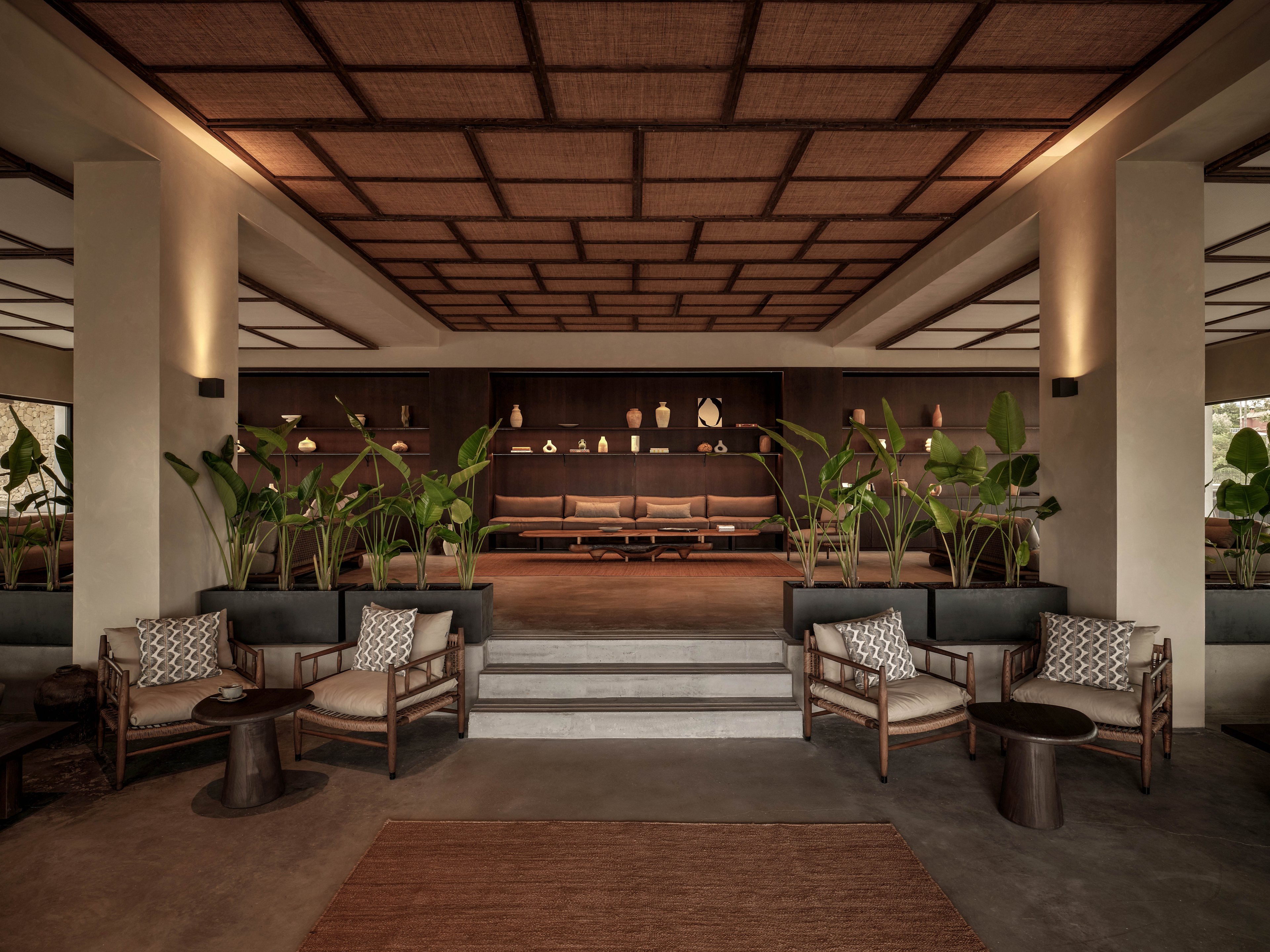 A contemporary lobby features symmetrical seating areas with cushioned chairs and sofas arranged around low tables. Large potted tropical plants line the central staircase, adding greenery to the neutral-toned interior. The ceiling showcases a geometric grid design, while built-in shelves at the back display decorative vases and objects. Warm, diffused lighting creates a calm and inviting atmosphere in the spacious lounge.