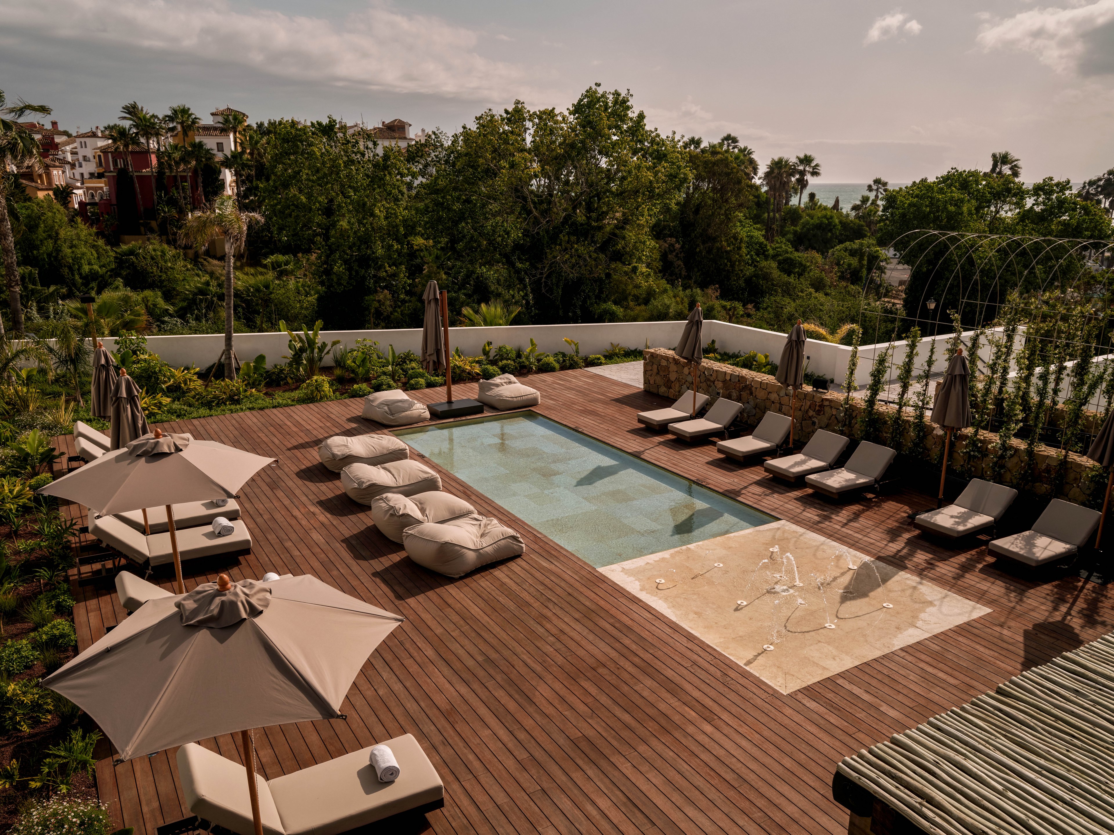 A contemporary rooftop pool area features a rectangular swimming pool surrounded by a wooden deck. Cushioned loungers and daybeds are arranged neatly along the sides, with large umbrellas providing shaded seating. Decorative fountains or water jets spray from a shallow section near the pool edge. Lush greenery and trees frame the space, while distant buildings and a hazy sky complete the tranquil resort-like setting.