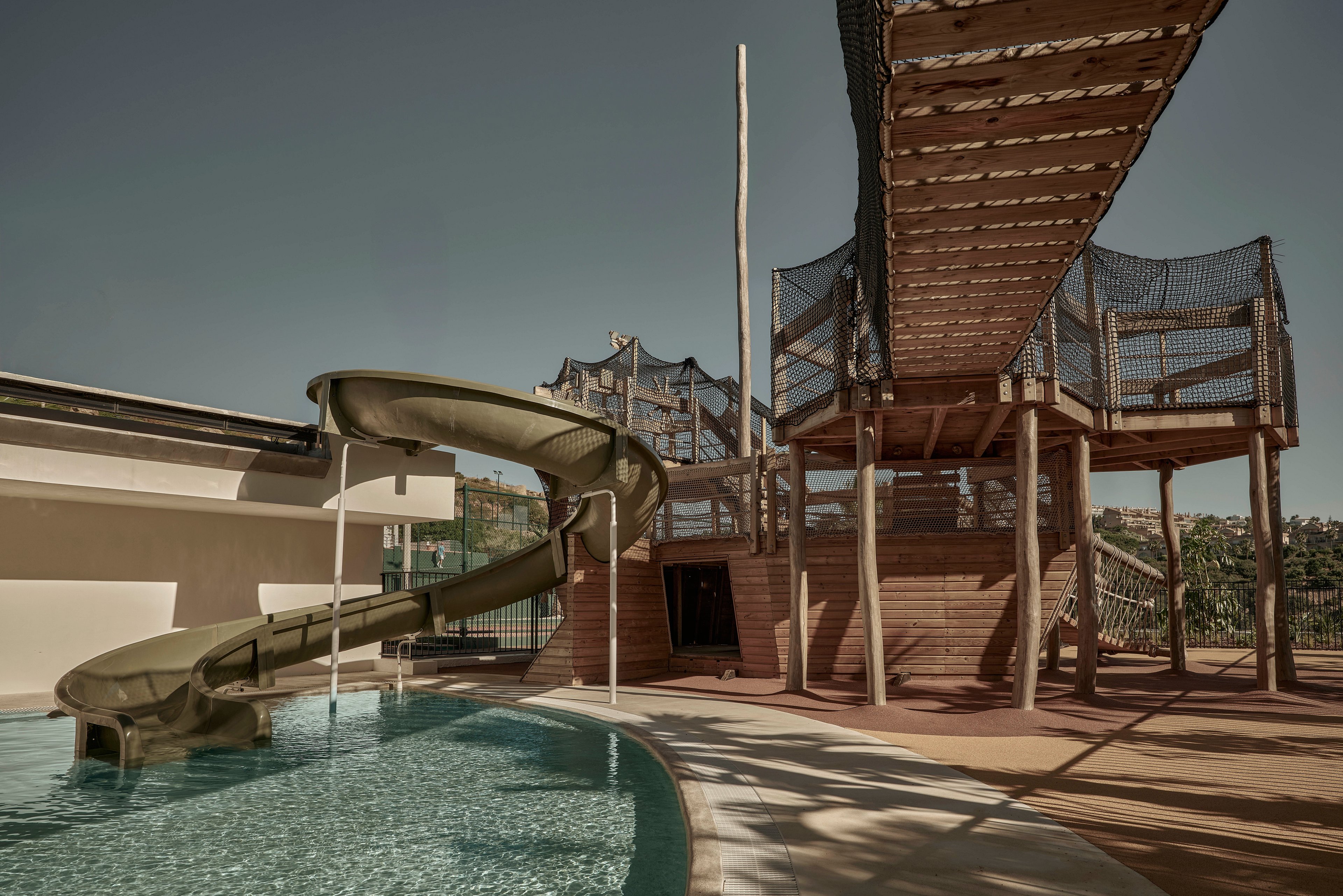 A curved waterslide ends in a shallow resort pool beside a wooden play structure. Above the pool, a high rope bridge and netted walkways create an elevated adventure course. The scene is empty of people, emphasizing the architecture and design of the recreational area. Warm, muted tones and strong sunlight give the image a calm, slightly nostalgic atmosphere.