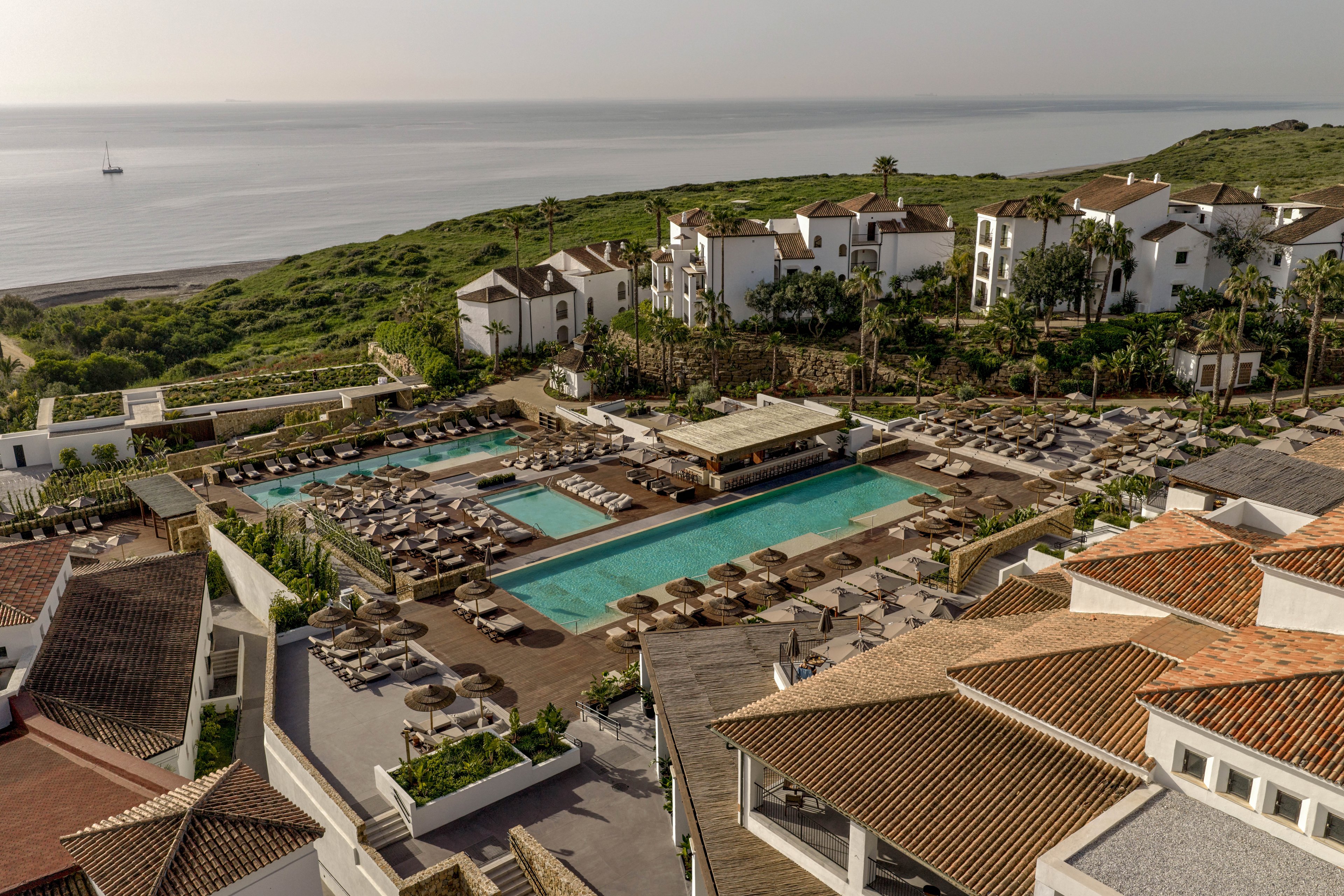 A large coastal resort features multiple swimming pools surrounded by rows of sun loungers and parasols. White Mediterranean-style buildings with terracotta roofs frame the pool terrace, creating a cohesive architectural look. The resort sits on a green hillside that gently slopes toward the shoreline and calm sea. In the distance, a small sailboat is visible on the water, enhancing the tranquil vacation atmosphere.