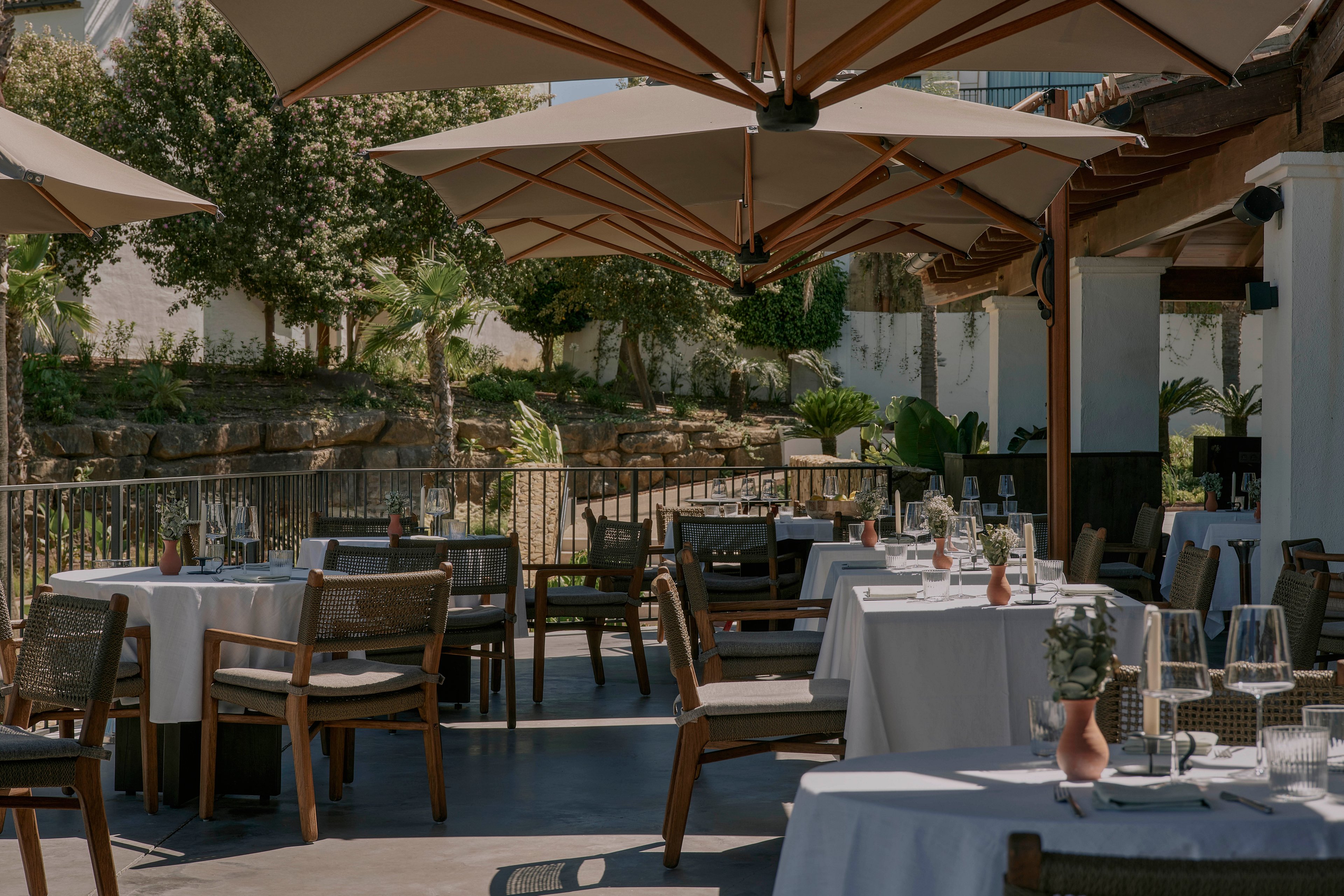 An upscale outdoor restaurant terrace is set with neatly arranged tables covered in white linens. Wooden chairs surround the tables, which are prepared with glassware, cutlery, and small decorative vases. Large beige umbrellas provide shade over the seating area, creating a comfortable dining environment. In the background, greenery and trees add a natural, tranquil atmosphere to the scene.