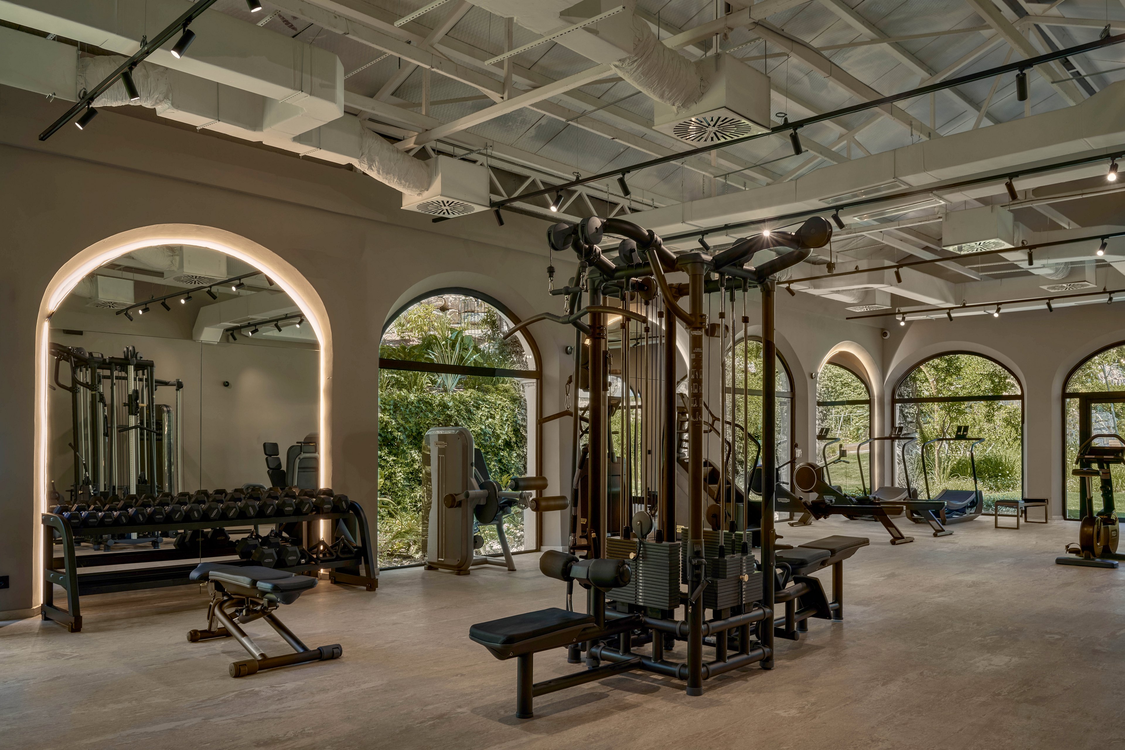 A spacious, modern gym interior features a variety of strength training machines and free weights. Large arched windows line the walls, allowing natural light to illuminate the workout area and reveal greenery outside. The neutral color palette and exposed ceiling create a clean, contemporary atmosphere. Benches, dumbbells, and cable machines are neatly arranged, suggesting a well-organized fitness space.