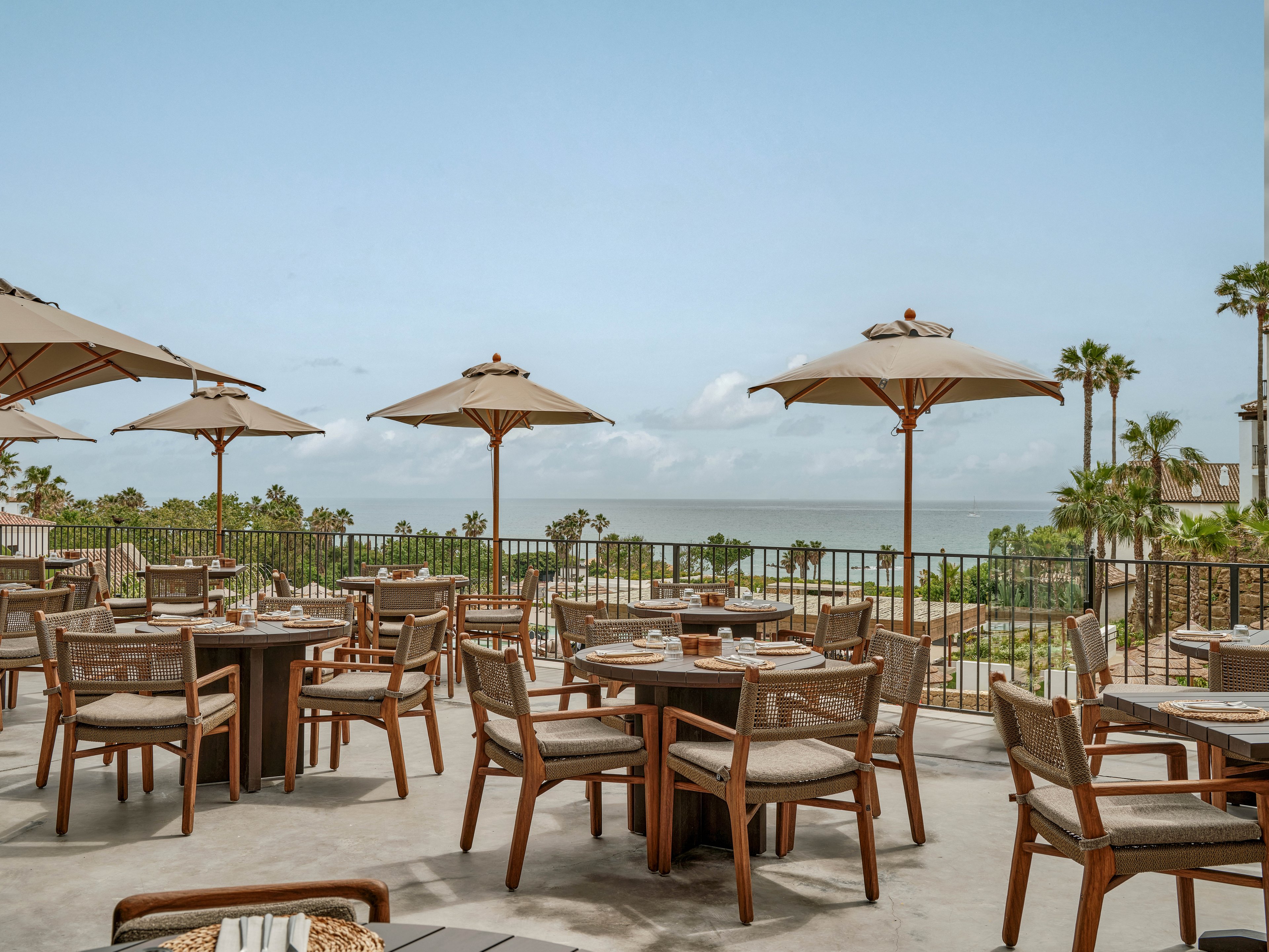 An open-air restaurant terrace features neatly arranged wooden tables and woven chairs under large beige umbrellas. The setting overlooks a coastal landscape with palm trees and a calm sea in the distance. Table settings with plates and glasses suggest a prepared dining experience. The overall atmosphere is relaxed and inviting, ideal for al fresco meals by the water.
