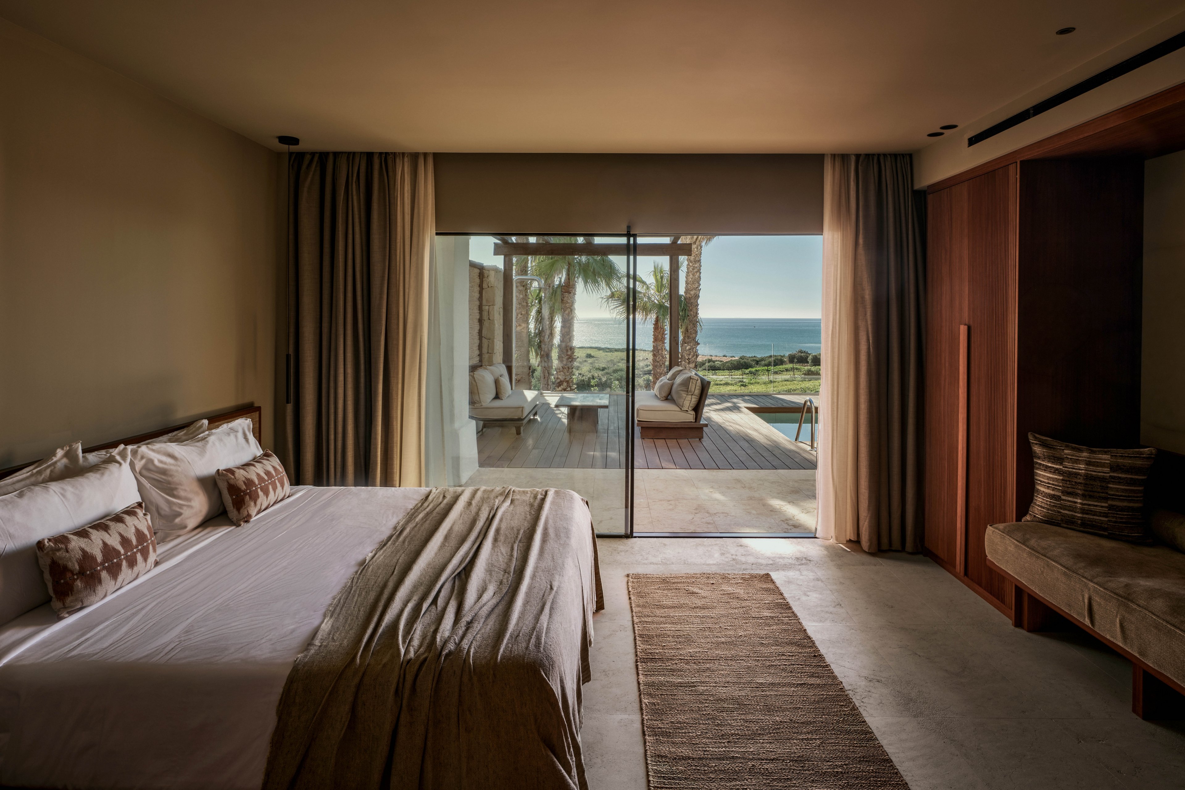 A contemporary bedroom features a large bed with neutral-toned linens and patterned pillows. Floor-to-ceiling glass doors open onto a terrace with lounge seating, palm trees, and a view of the ocean in the distance. Warm lighting and soft curtains create a cozy, tranquil atmosphere. Minimalist furniture and earthy colors emphasize a luxurious, relaxing resort setting.