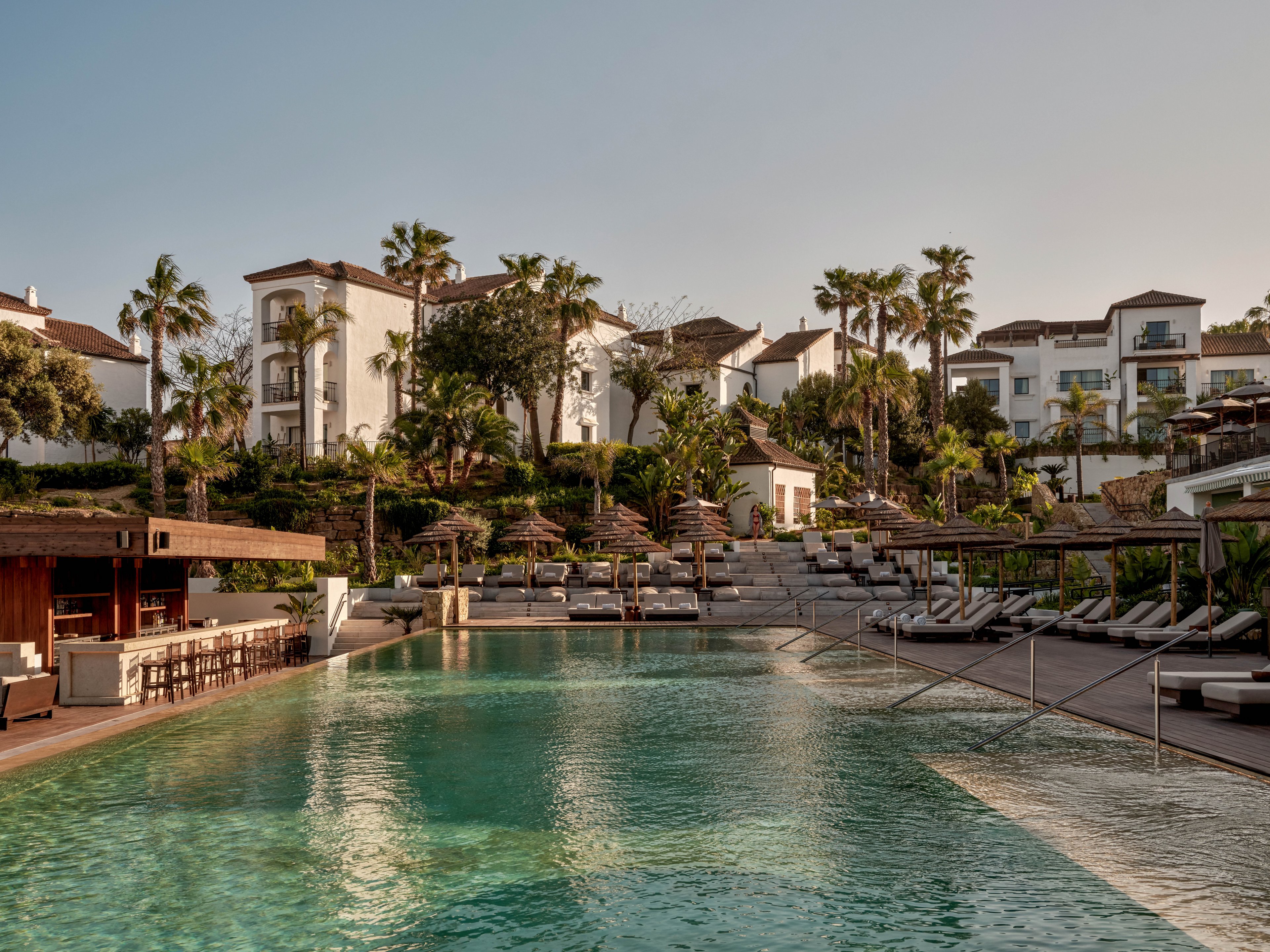 A large, tranquil swimming pool sits at the center of a luxury hillside resort. White multi-story buildings with terracotta roofs rise behind the pool, surrounded by lush greenery and tall palm trees. Numerous sun loungers and umbrellas line the poolside terraces, creating a relaxed vacation atmosphere. Warm, late-afternoon light casts soft reflections on the calm water, enhancing the serene and upscale setting.