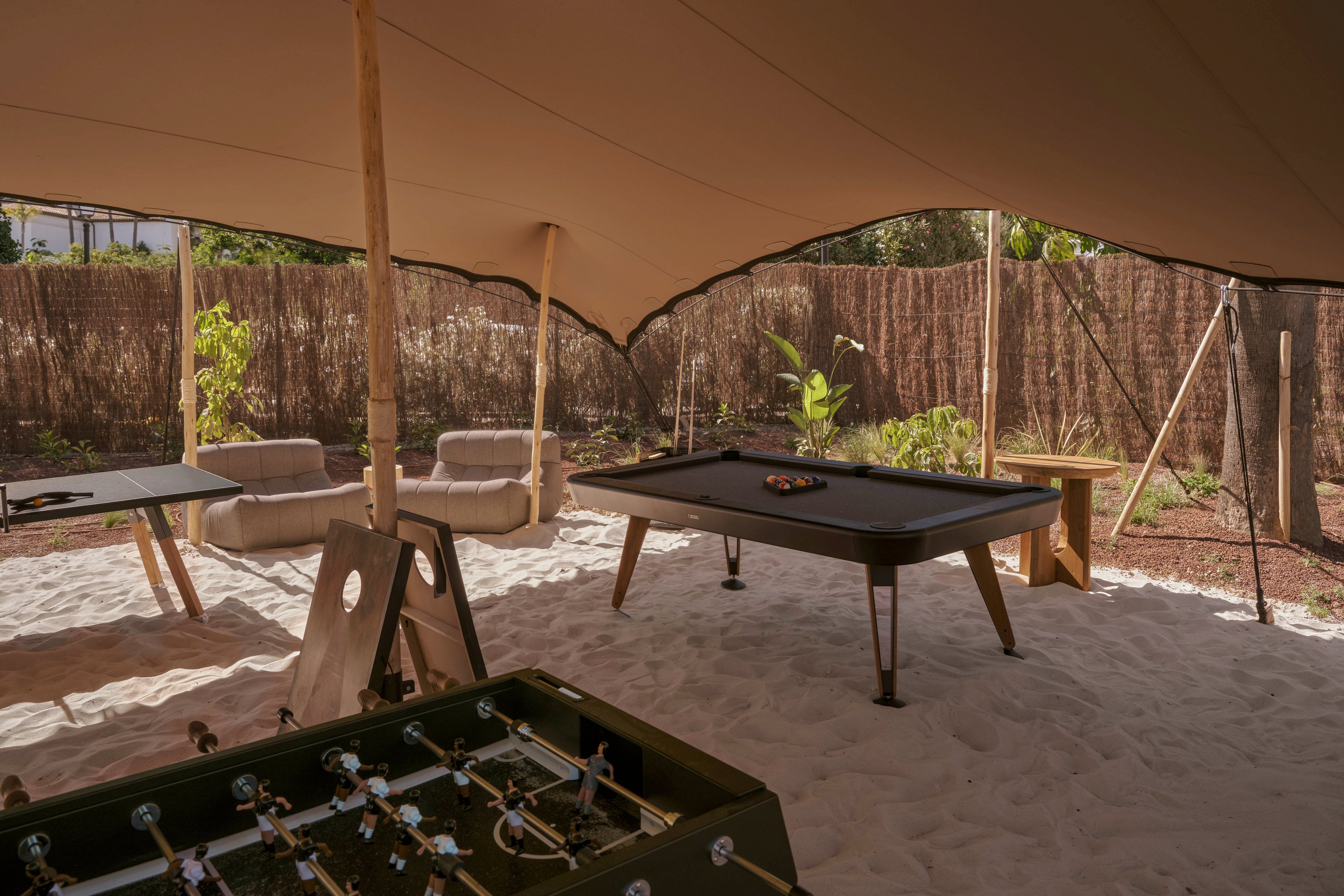 An outdoor recreation area features a pool table set on a sandy surface under a large canopy. A foosball table stands in the foreground, while a side table and additional seating are arranged nearby. The space is enclosed by a natural fence with plants and trees providing a relaxed, shaded atmosphere. Soft lounge chairs and warm lighting contribute to a casual, resort-like setting.