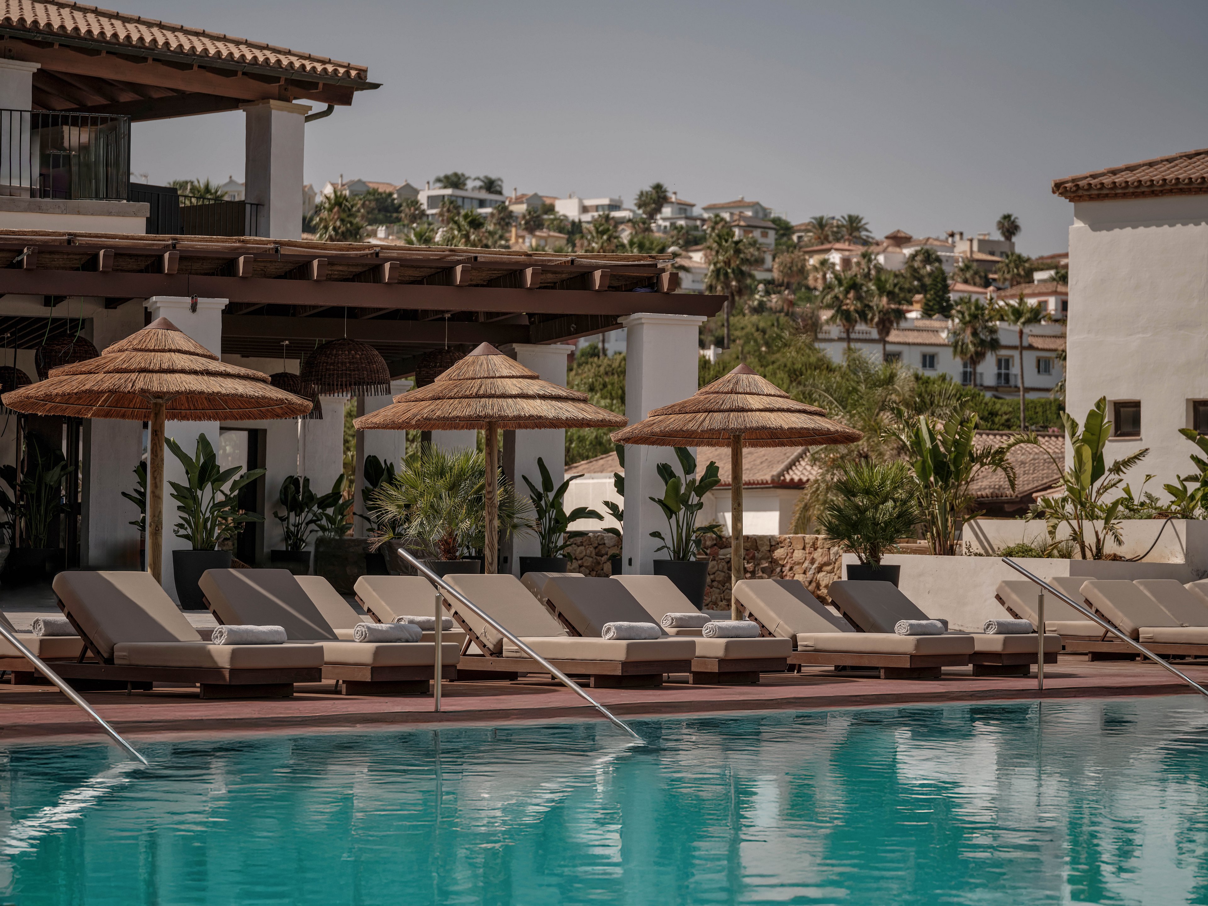 A serene resort swimming pool is lined with cushioned sun loungers and straw parasols. The scene is set in a warm, sunny climate with clear blue water in the foreground. White stucco buildings and terraces with wooden pergolas frame the pool area. In the background, a hillside dotted with houses and palm trees adds to the relaxed vacation atmosphere.