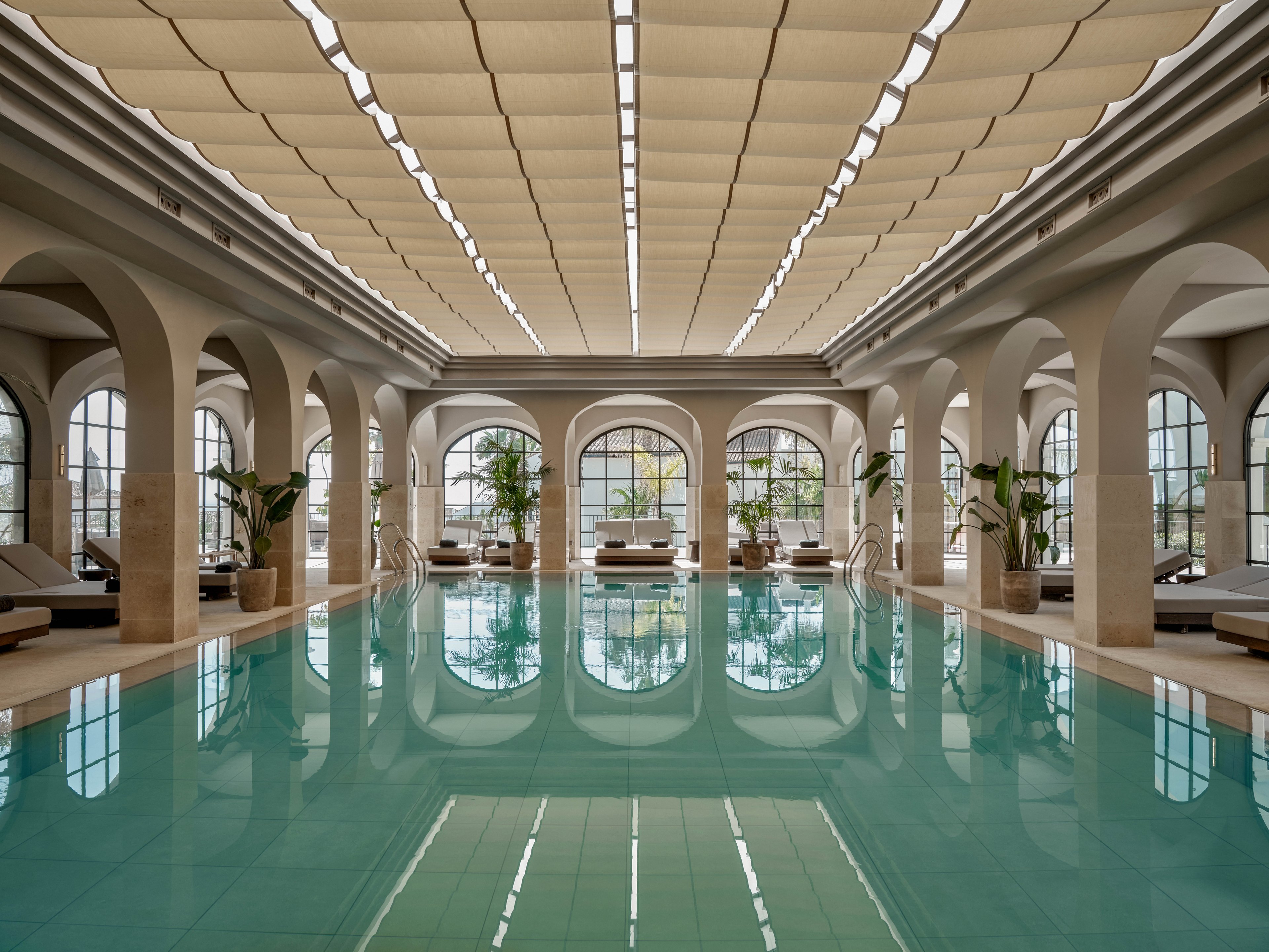 A serene indoor swimming pool is framed by elegant arches and large arched windows. Soft natural light filters through the ceiling panels, reflecting gently on the turquoise water. Lounge chairs and potted plants line the sides, creating a relaxing spa-like atmosphere. The symmetrical architecture and muted color palette emphasize calm and sophistication.