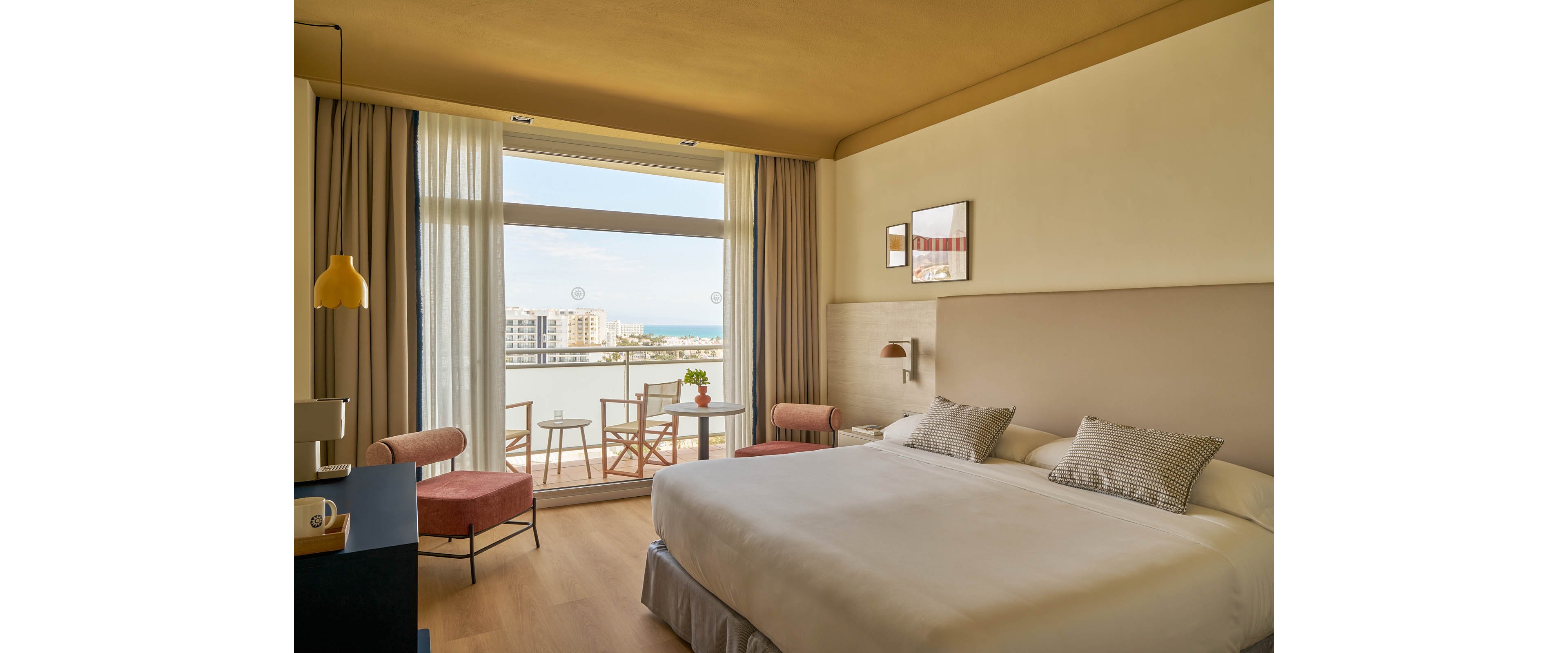A spacious, contemporary hotel room features a large bed with neutral bedding and accent pillows. The room opens to a balcony with two chairs and a table, overlooking a cityscape and distant water. Soft natural light fills the space, highlighting minimalist decor, wooden flooring, and warm tones. No visible text or numeric values are present in the image.