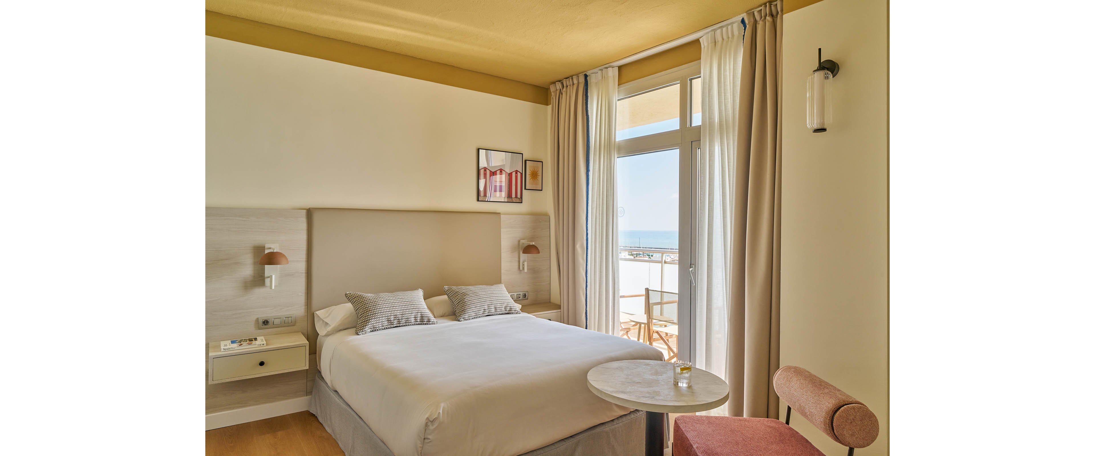 A sunlit hotel room features a neatly made double bed with neutral bedding and two pillows. The space includes a small round table, a pink upholstered chair, and wall-mounted bedside lamps. Large windows with sheer curtains open to a balcony overlooking the sea, creating an airy and inviting atmosphere. Minimalist decor and soft beige tones enhance the tranquil mood.
