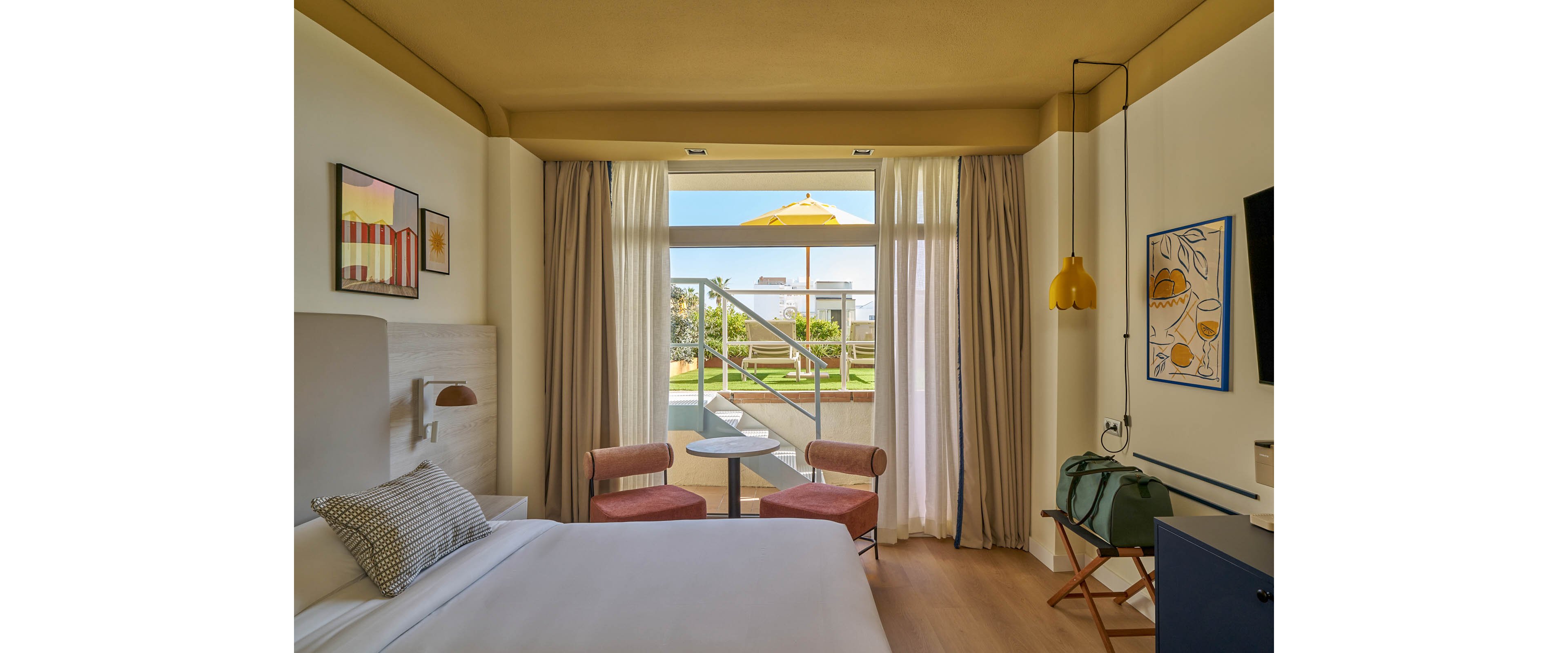 A contemporary hotel room features a neatly made bed, two pink chairs, and a small table near a large window. The window opens to a patio with a yellow umbrella and green garden area visible outside. The room is decorated with framed artwork, a hanging yellow lamp, and a travel bag on a wooden chair, creating a welcoming and relaxed atmosphere.
