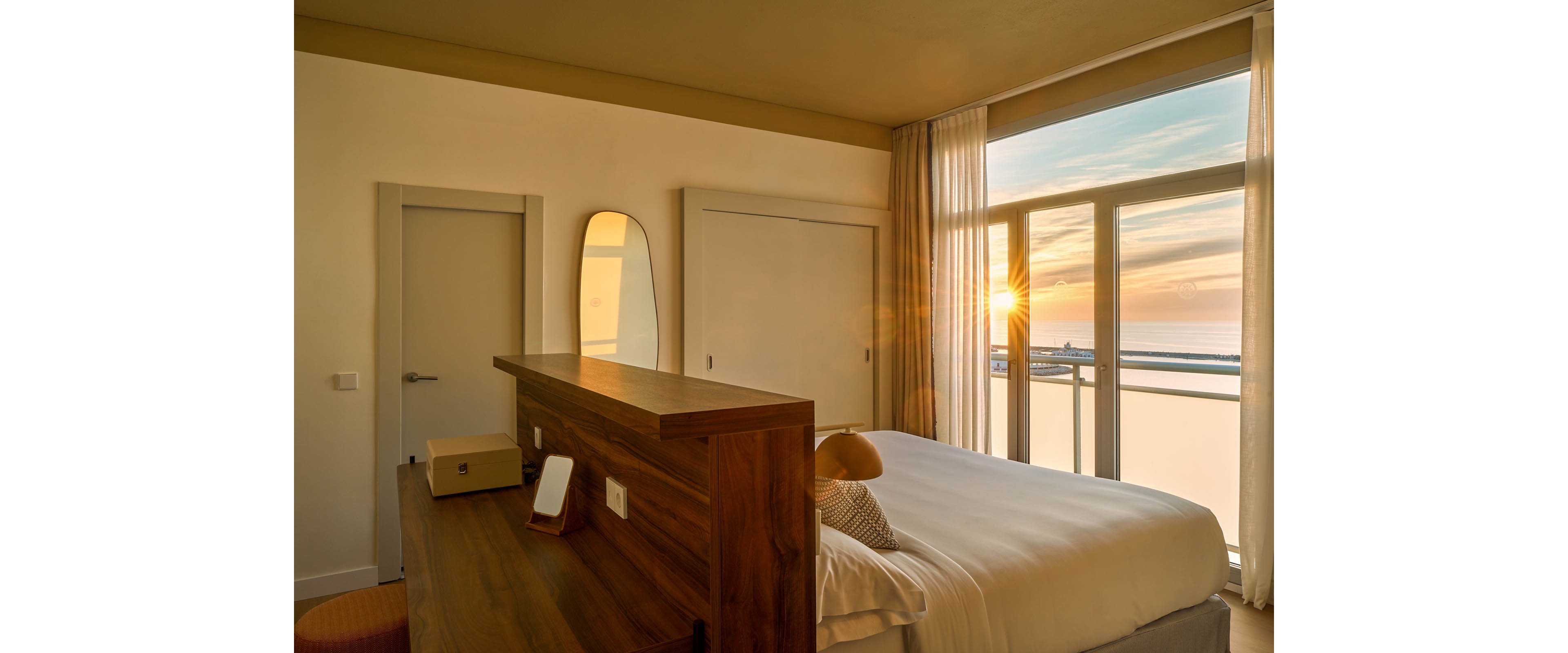 A contemporary hotel room features a neatly made bed, wooden furniture, and a large window overlooking the ocean at sunset. Warm sunlight streams through sheer curtains, casting a golden glow across the room. Minimalist decor includes a mirror, lamp, and small storage box, creating a calm and inviting atmosphere.