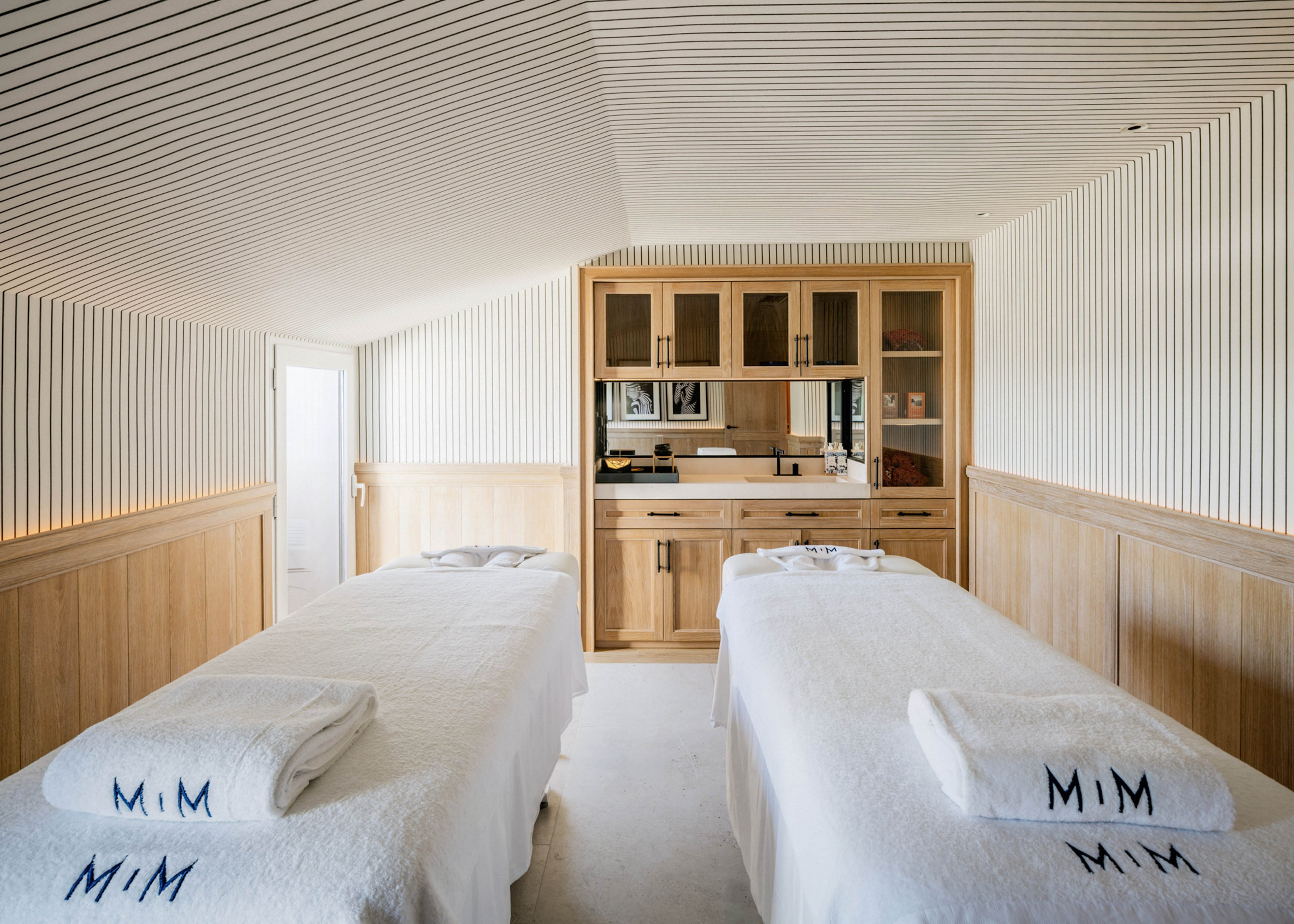 A serene spa treatment room features two massage tables covered in white linens, each with a towel embroidered with 'MiM'. The space is designed with light wood paneling and cabinetry, creating a calm and inviting atmosphere. The room is well-lit with natural and ambient lighting, and the overall palette is neutral and soothing. No people are present, emphasizing tranquility and relaxation.