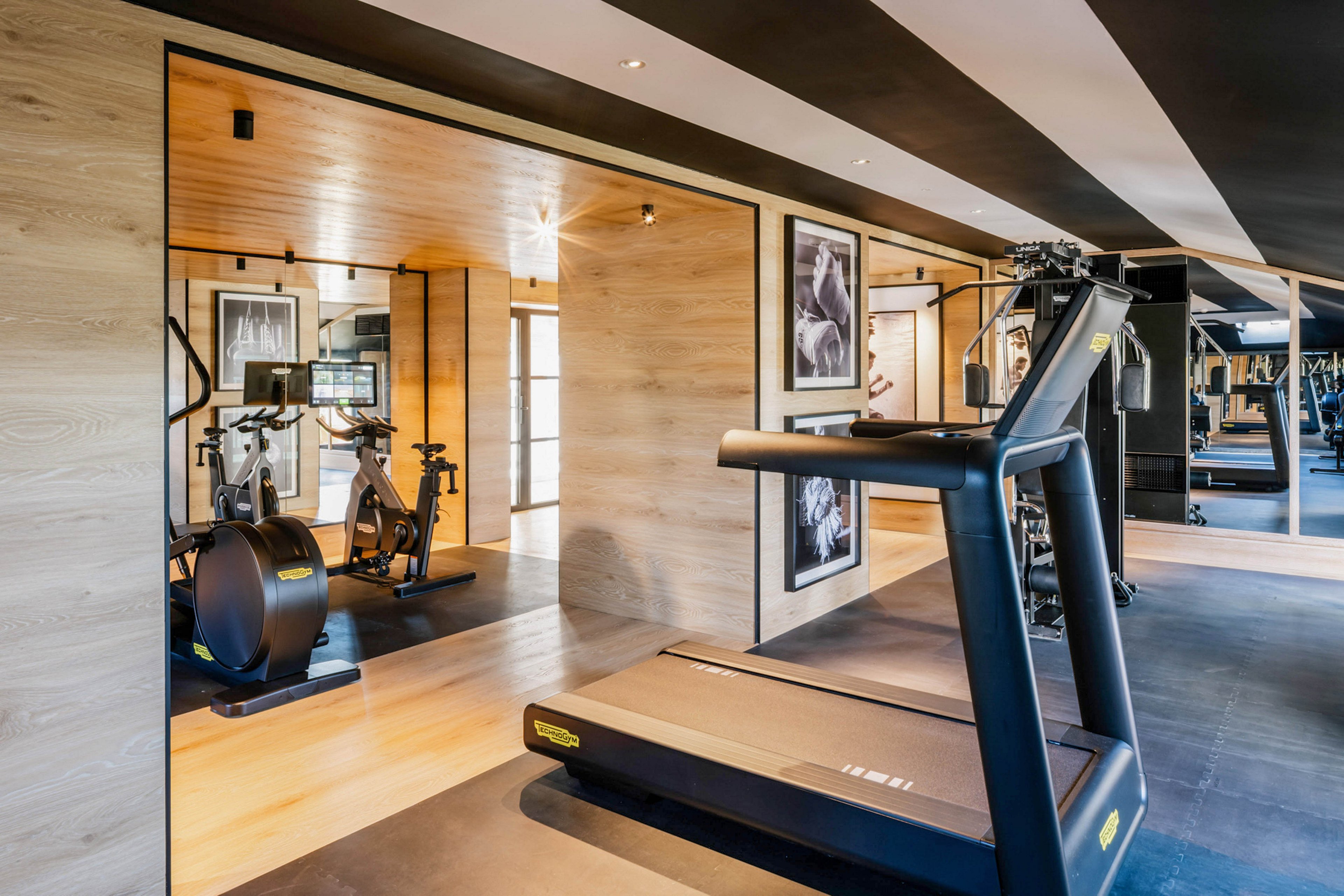 A contemporary fitness center features a treadmill in the foreground and stationary exercise bikes in a separate room. The space is designed with light wood paneling, large mirrors, and framed black-and-white photographs on the walls. The gym is well-lit with natural and artificial lighting, creating a clean and inviting atmosphere. No visible people or numeric values are present in the image.