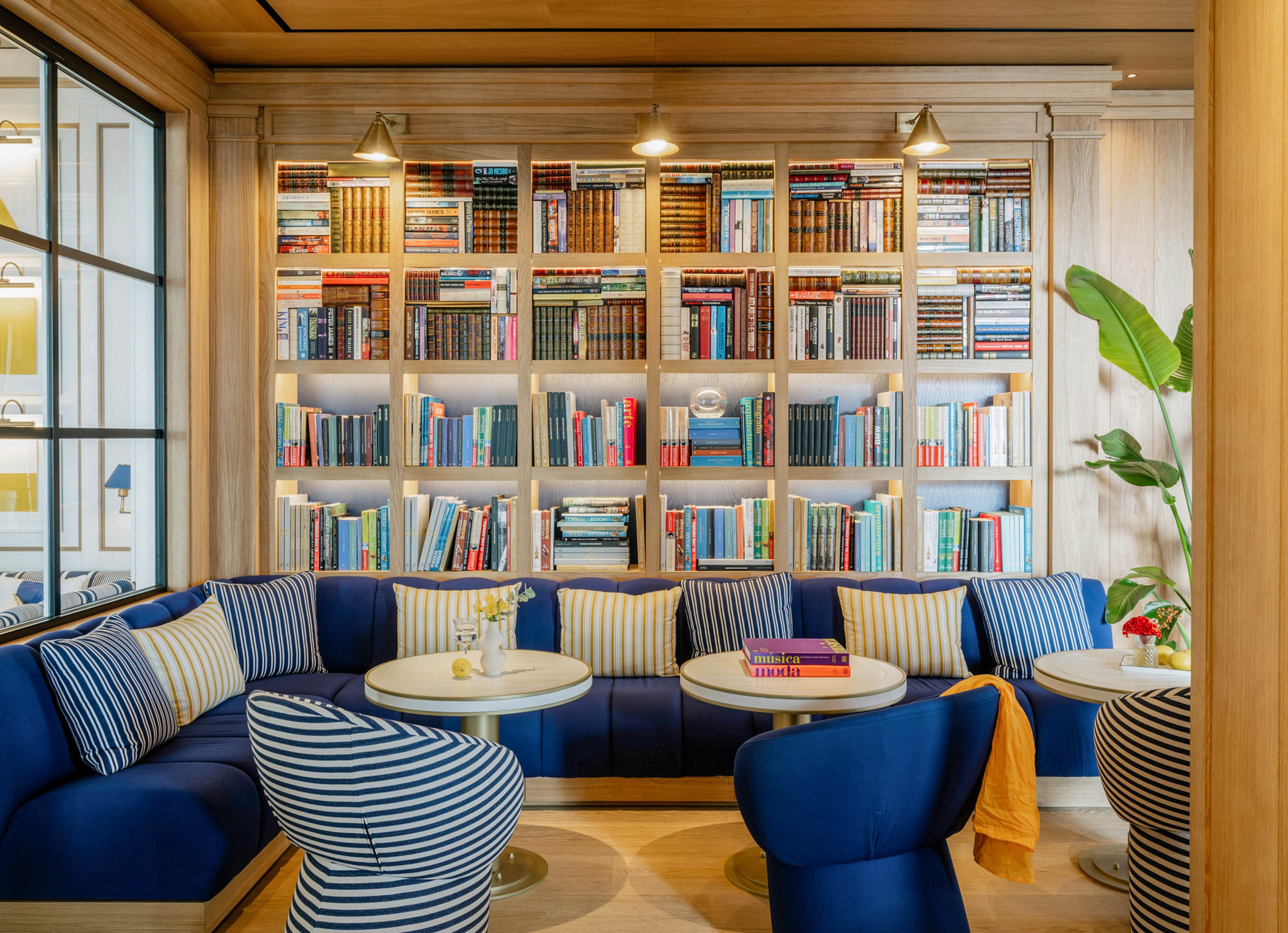A stylish lounge area features deep blue cushioned seating with striped pillows arranged around small round tables. The backdrop is a large bookshelf filled with colorful books, illuminated by overhead lights. The setting is bright and inviting, with a mix of wood tones, greenery, and a relaxed, contemporary atmosphere. No visible text or numbers are present in the image.