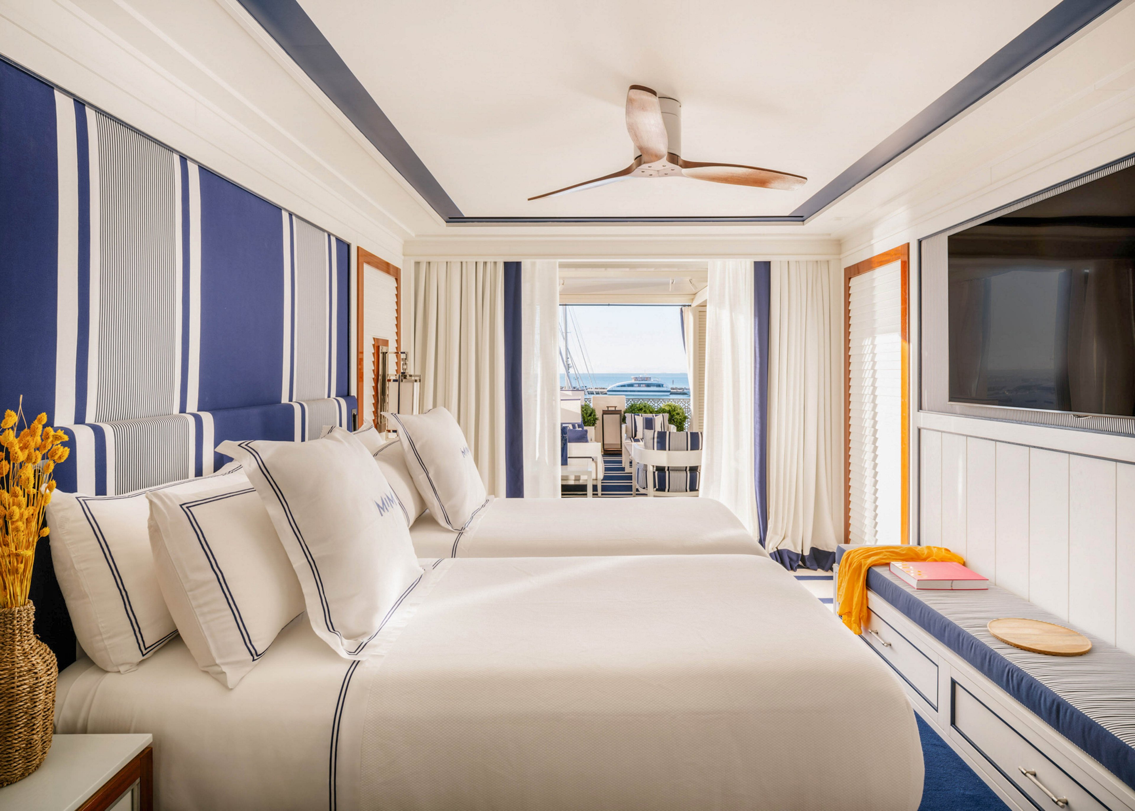 A stylish hotel room features two neatly made twin beds with white and navy blue linens. The space is decorated in a nautical theme with bold striped walls, a ceiling fan, and a large wall-mounted television. Natural light fills the room from a balcony that overlooks the ocean, creating a fresh and inviting atmosphere. Accents include a bench with a yellow towel and a vase of yellow flowers, enhancing the coastal vibe.