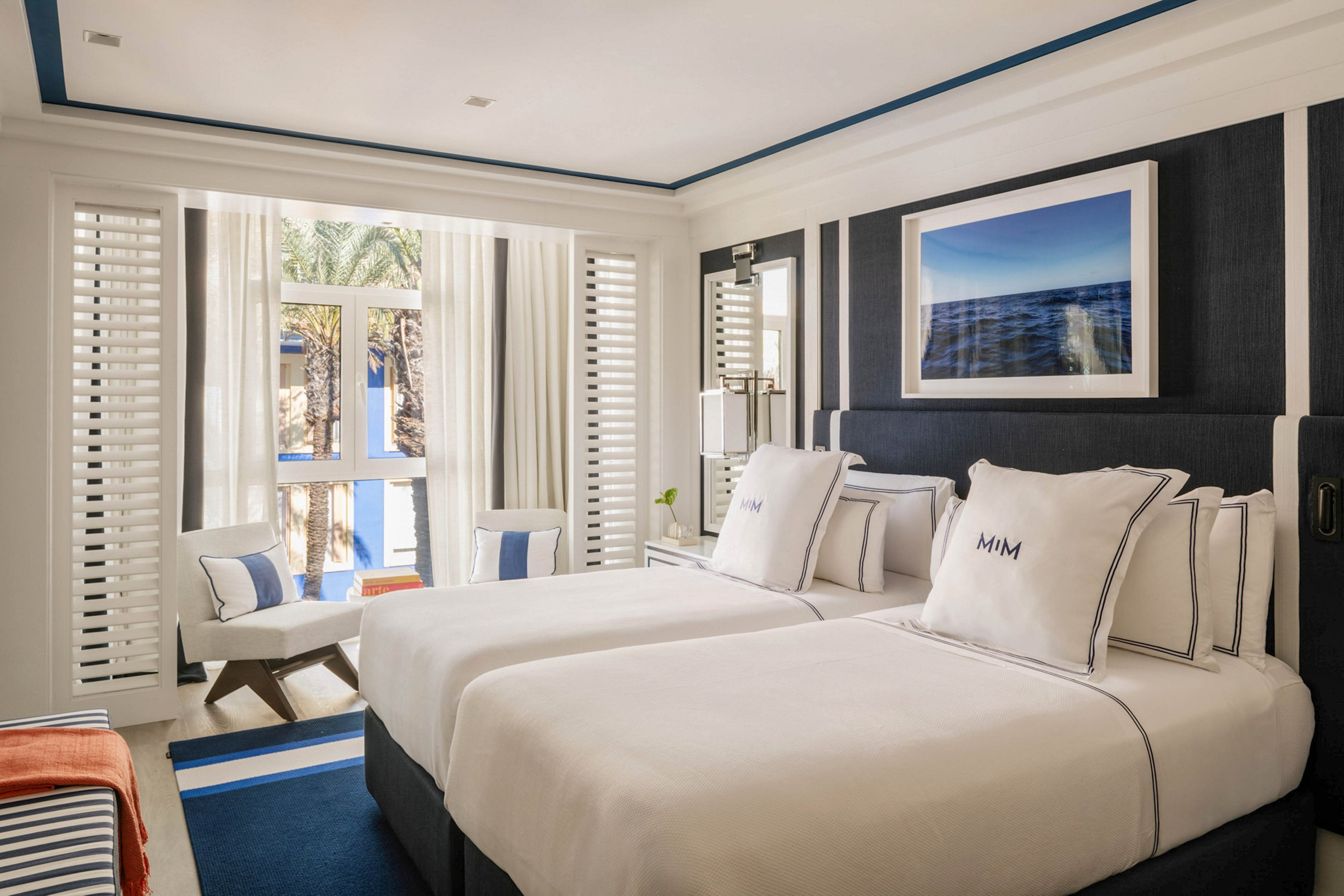 A stylish hotel room features two neatly made twin beds with white linens and navy trim. The space is filled with natural light from a large window, which is framed by white shutters and curtains, offering a view of palm trees outside. The decor is modern and nautical, with blue and white accents, a framed seascape photograph, and a cozy seating area. The pillows display the text 'MIM' clearly. The overall mood is fresh, inviting, and tranquil.