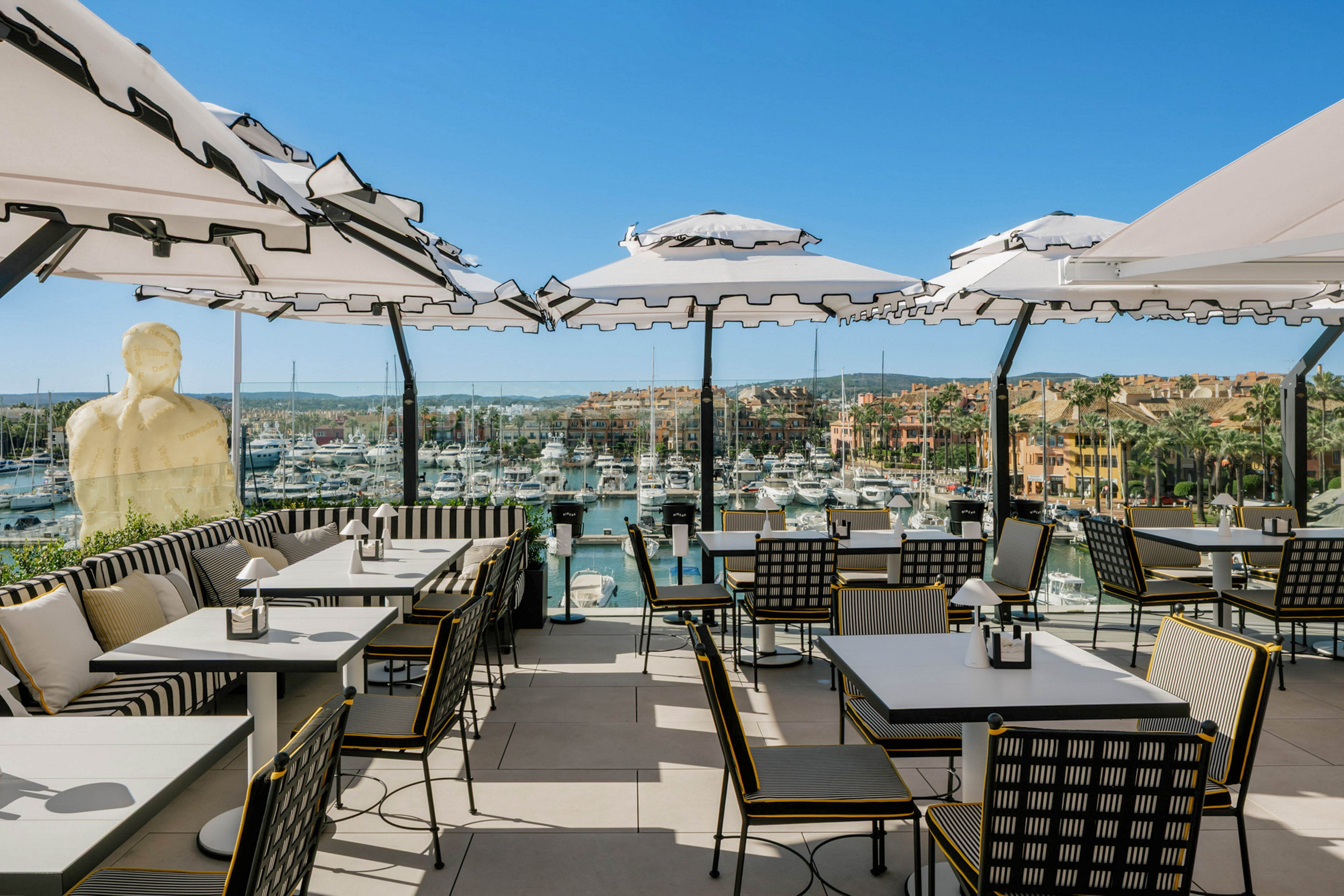 A stylish outdoor restaurant terrace overlooks a marina filled with boats and yachts under a clear blue sky. The seating area features modern tables, striped cushions, and large white umbrellas providing shade. The setting is bright and inviting, with a relaxed, upscale atmosphere and a scenic waterfront view.