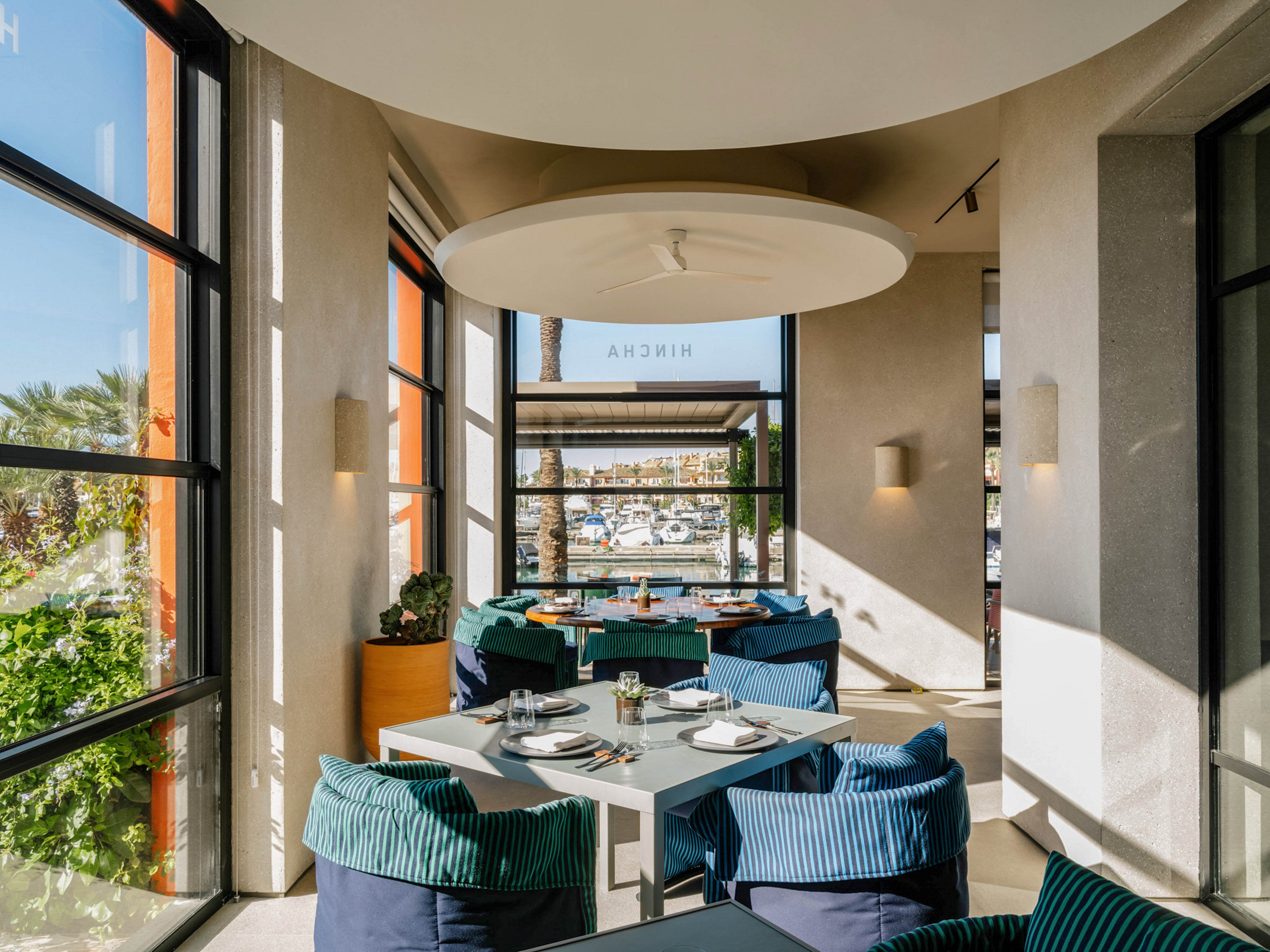 A contemporary restaurant dining area is bathed in natural sunlight, featuring tables set for service and plush blue seating. Large windows offer a scenic view of a marina with boats docked outside. The space is decorated in a minimalist style with neutral tones and clean lines, creating a relaxed and inviting atmosphere. The word 'NIKSHA' is visible on the glass, possibly indicating the venue name.