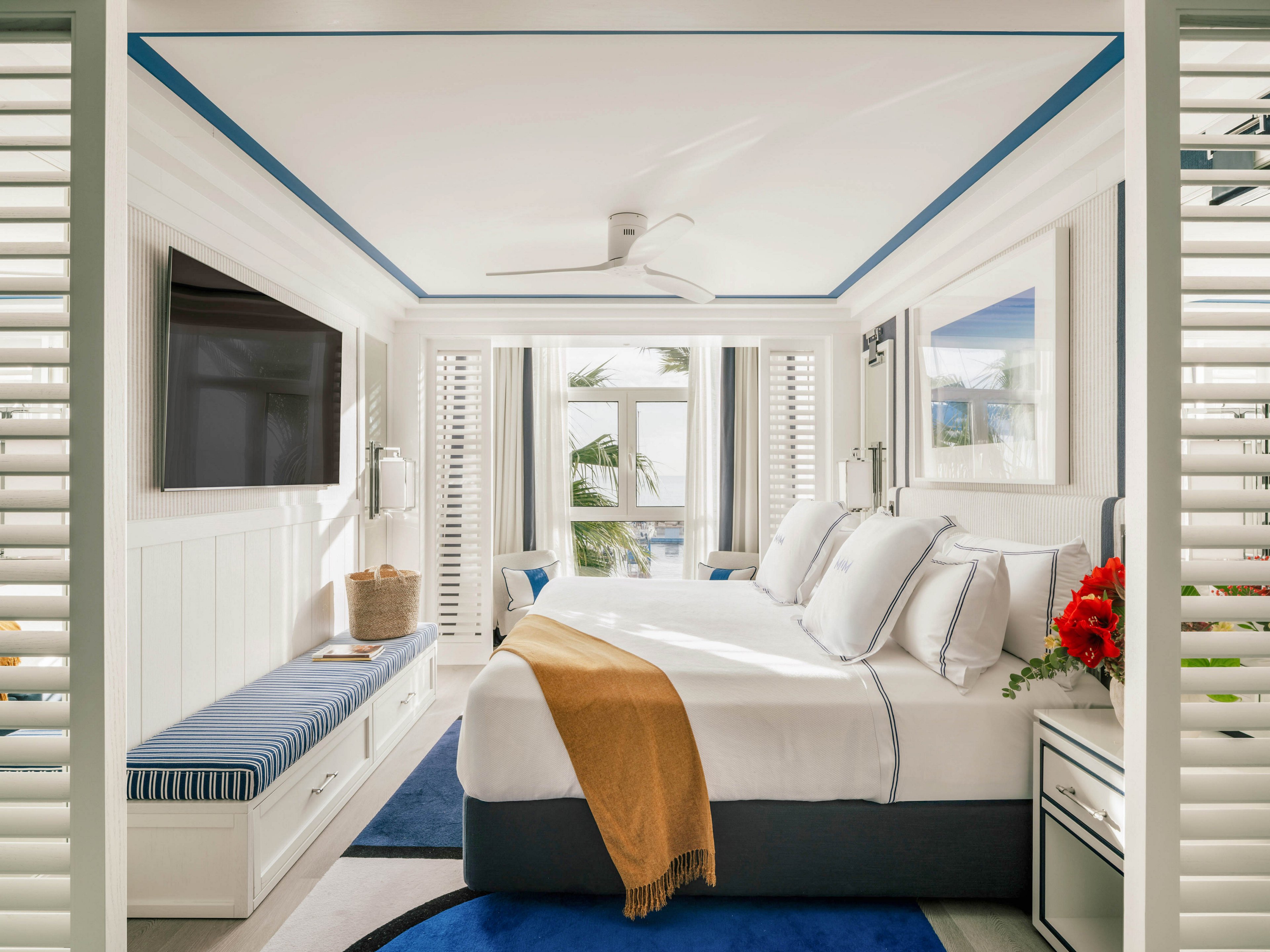 A sunlit bedroom features a large bed with white and blue linens, accented by a mustard throw. The room includes a wall-mounted flat-screen TV, a built-in bench with striped cushion, and a bouquet of red flowers on the nightstand. Large windows reveal palm trees and an ocean view, creating a fresh, airy atmosphere with coastal-inspired decor. The palette is dominated by white, blue, and natural tones.