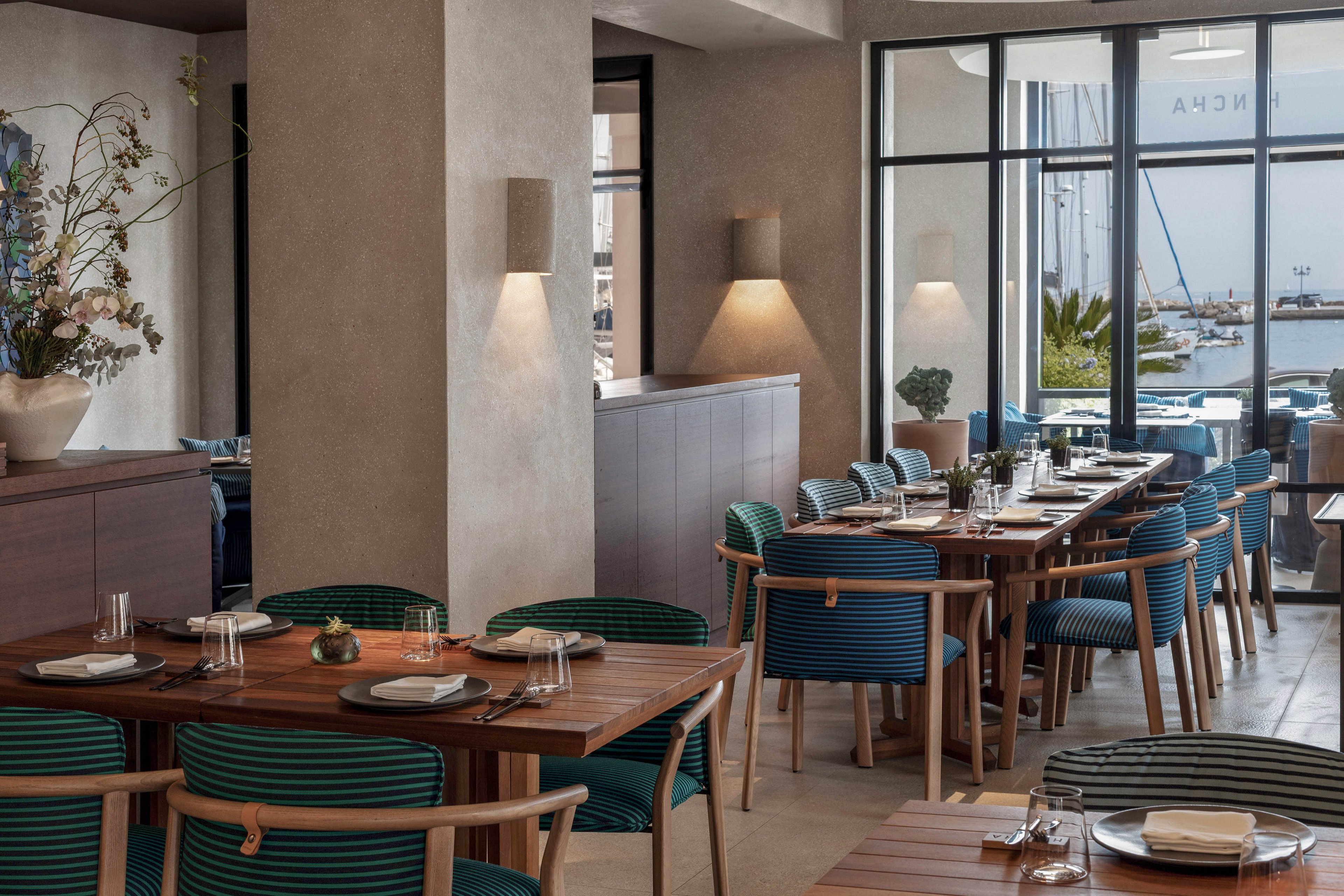A contemporary restaurant dining area features neatly arranged wooden tables and blue upholstered chairs. The setting is bright and airy, with large windows offering a view of a marina and docked boats outside. Table settings include plates, glasses, and cutlery, while neutral walls and minimalist decor create a calm, inviting atmosphere. No visible text or numbers are present in the image.