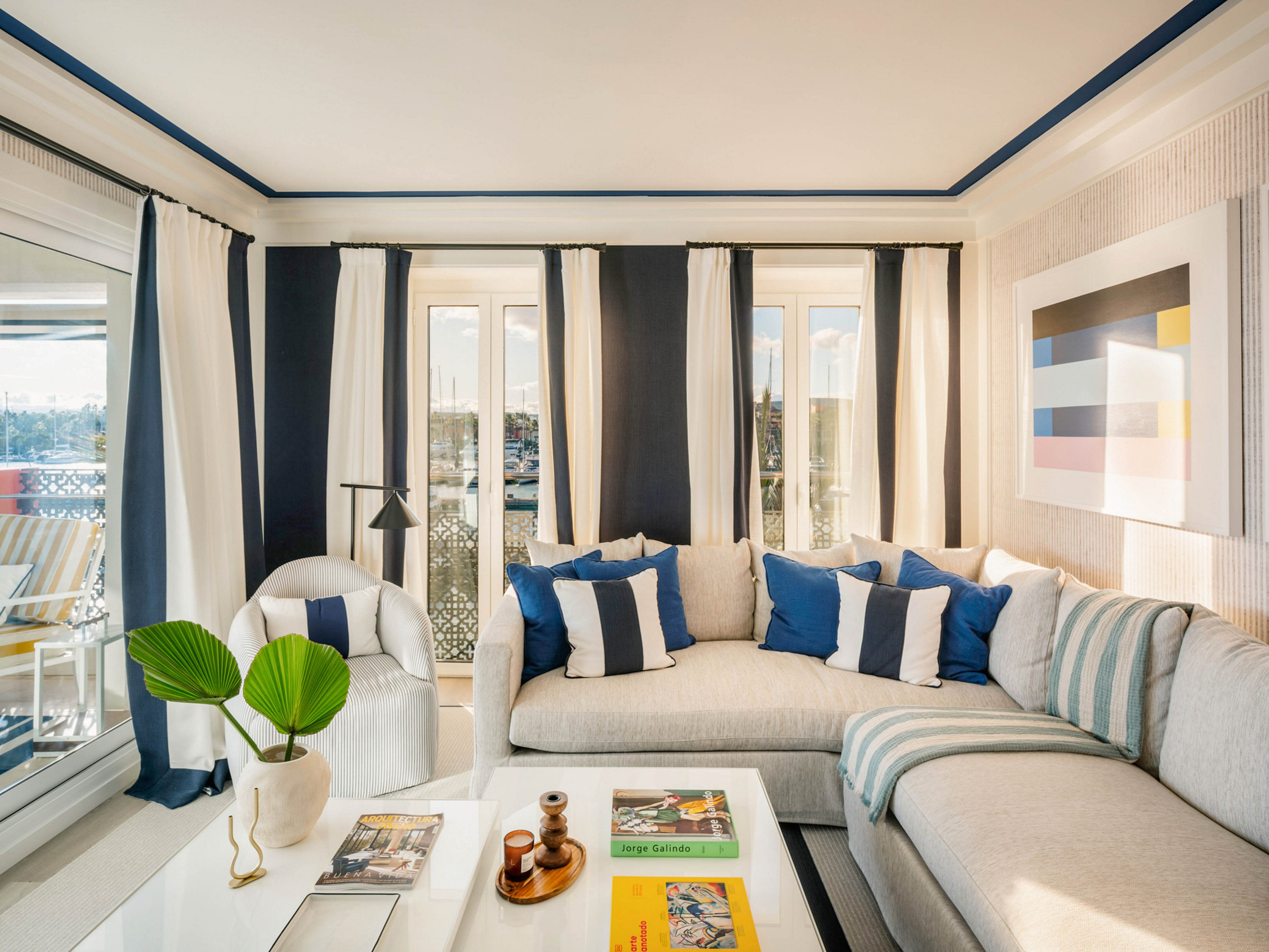 A contemporary living room features a light sectional sofa with blue and white throw pillows, set against large windows with matching curtains. The space is decorated with a geometric wall art piece, a white coffee table holding books and a vase with green leaves, and a cozy armchair. Sunlight fills the room, highlighting the clean lines and nautical-inspired color palette. The overall mood is bright, inviting, and stylish.