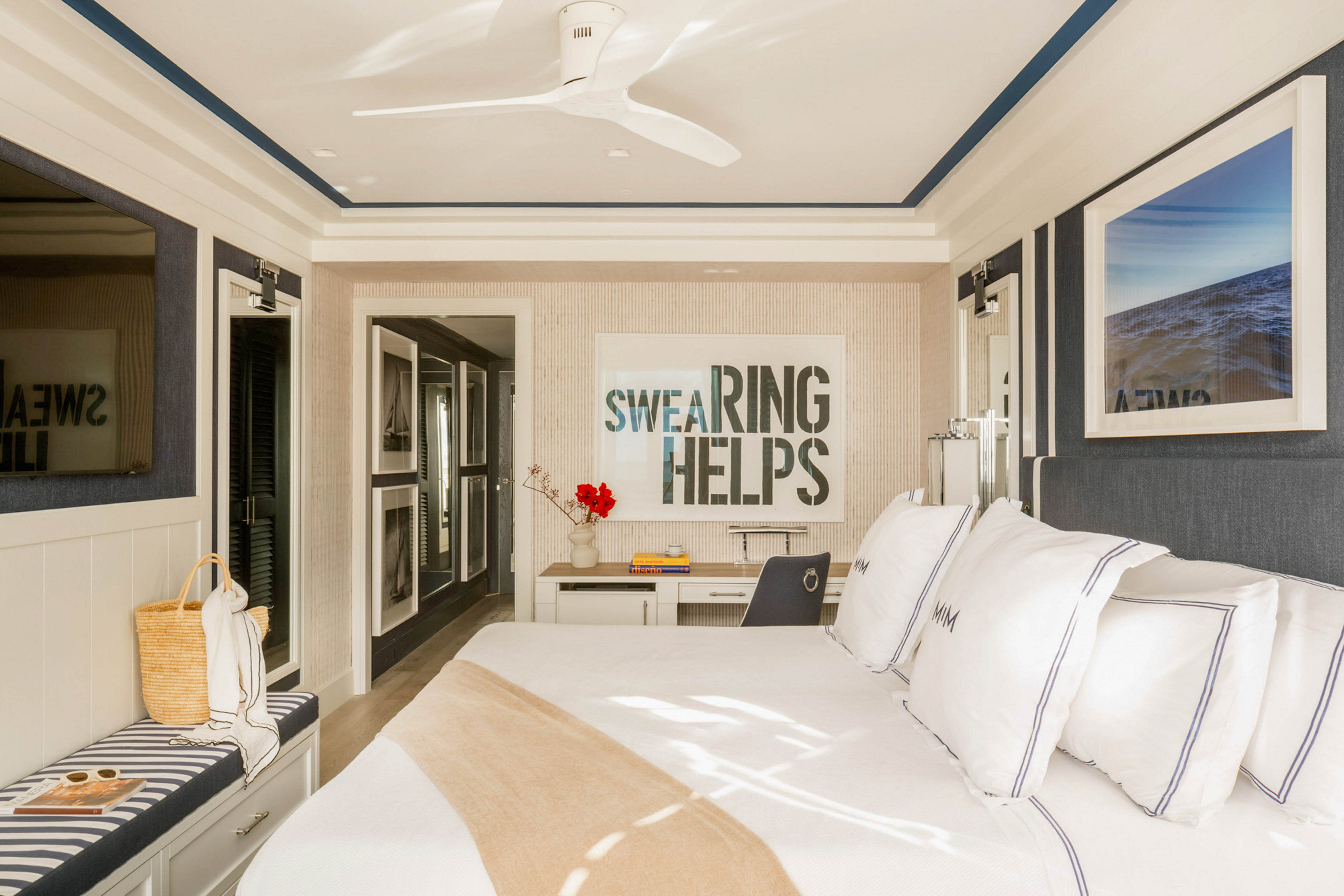 A contemporary hotel room features a neatly made bed with white linens and accent pillows, a striped bench, and a workspace. The setting is bright and inviting, with natural light streaming in and a large framed artwork on the wall displaying the text 'SWEARING HELPS.' The decor includes neutral tones with navy accents, a ceiling fan, and modern furnishings, creating a relaxed and sophisticated atmosphere.