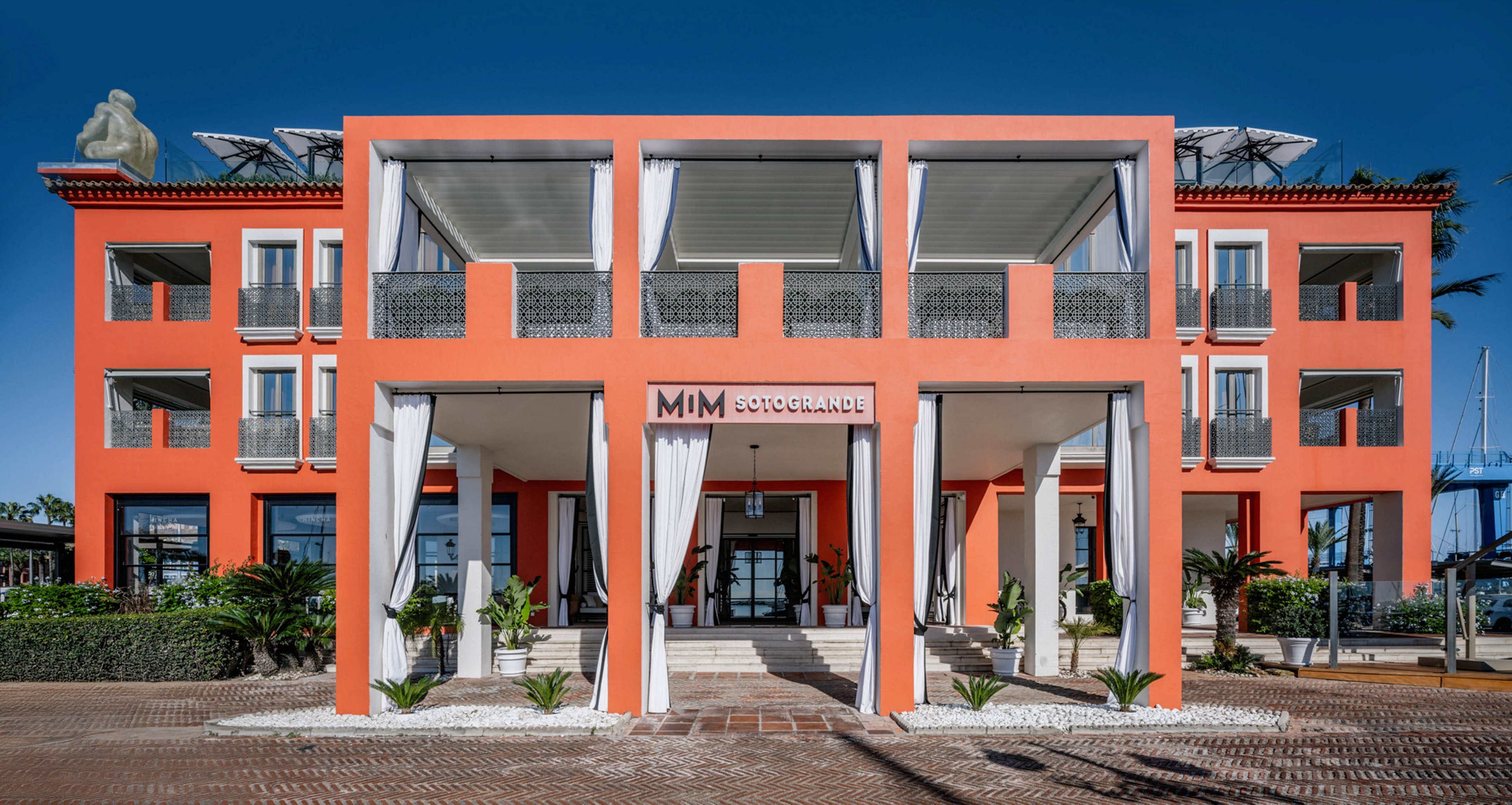 A contemporary hotel building features a bold coral exterior with white architectural accents and balconies. The entrance is framed by tall columns and draped curtains, with tropical plants lining the walkway. The sign above the entrance reads 'MIM Sotogrande,' clearly identifying the property. The sky is clear, enhancing the vibrant color palette and inviting atmosphere.