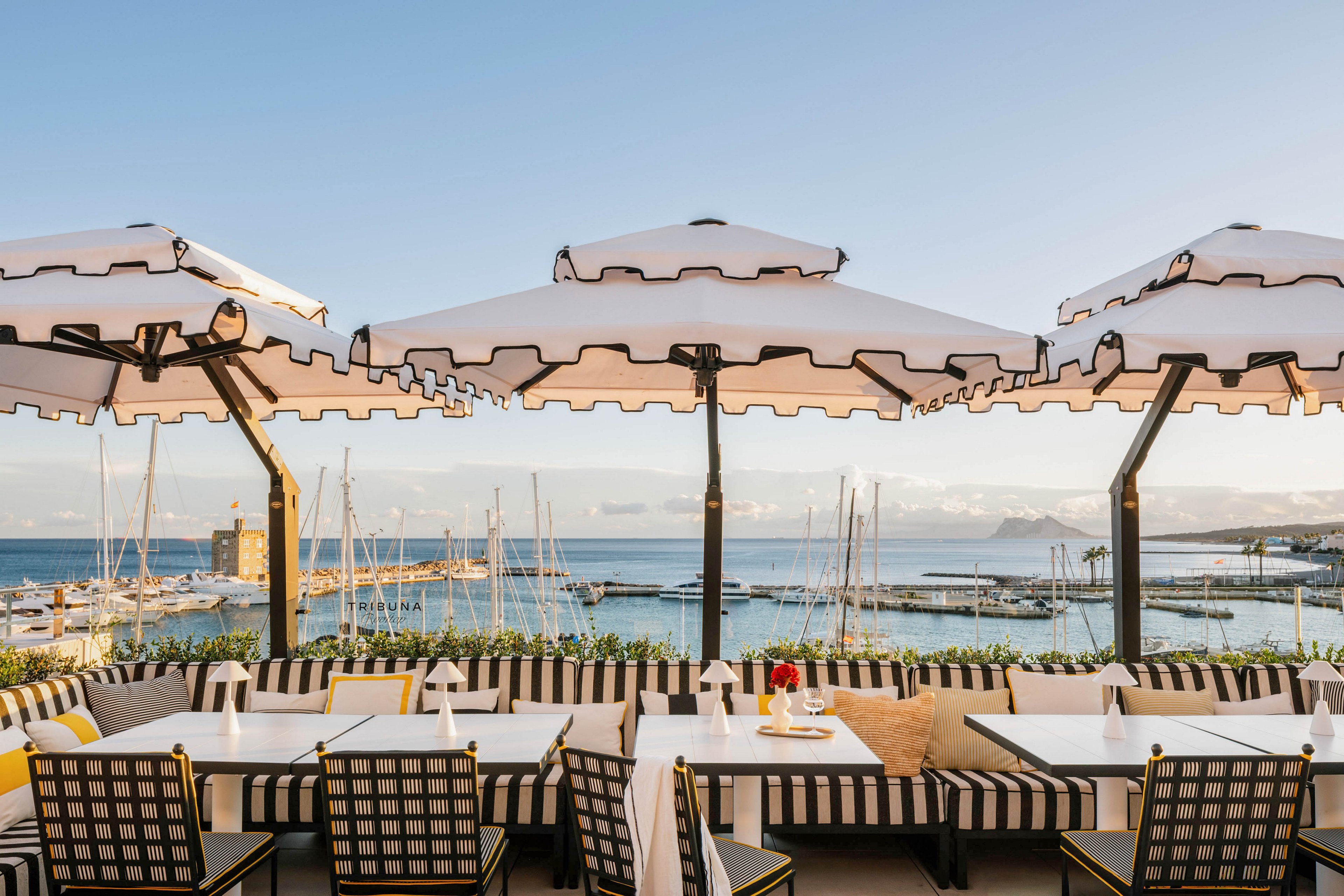 A stylish outdoor restaurant terrace overlooks a marina filled with sailboats and yachts. The seating area features black and white striped cushions and matching large umbrellas, creating a chic and inviting atmosphere. The tables are set for dining, and the scene is bathed in warm, golden sunset light. The calm sea and distant mountains enhance the relaxed, upscale mood.