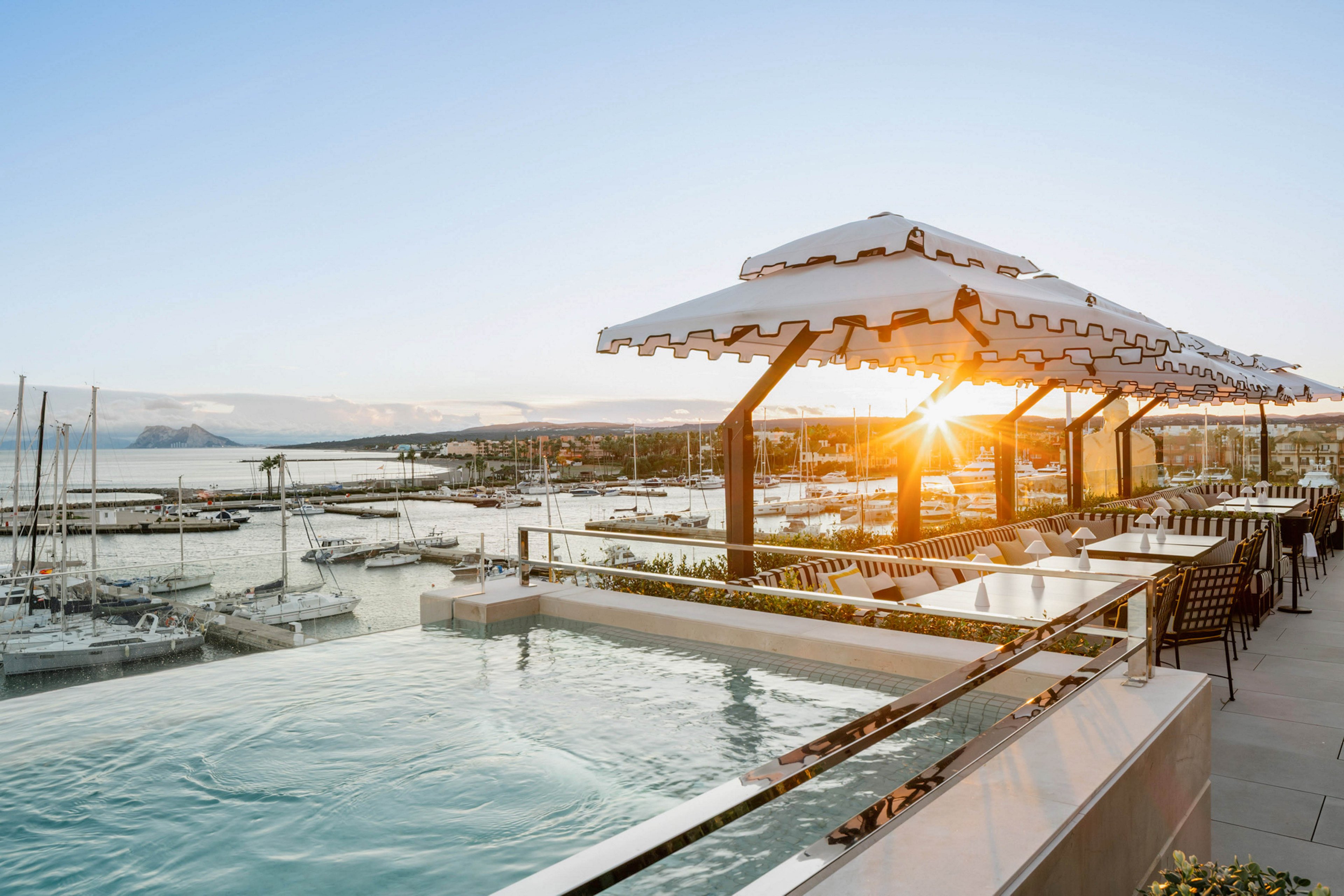 A luxurious rooftop infinity pool offers a stunning view of a marina filled with docked sailboats and yachts. The setting sun creates a warm, golden glow across the water and modern outdoor seating area. Striped awnings provide shade, enhancing the relaxed and upscale atmosphere.